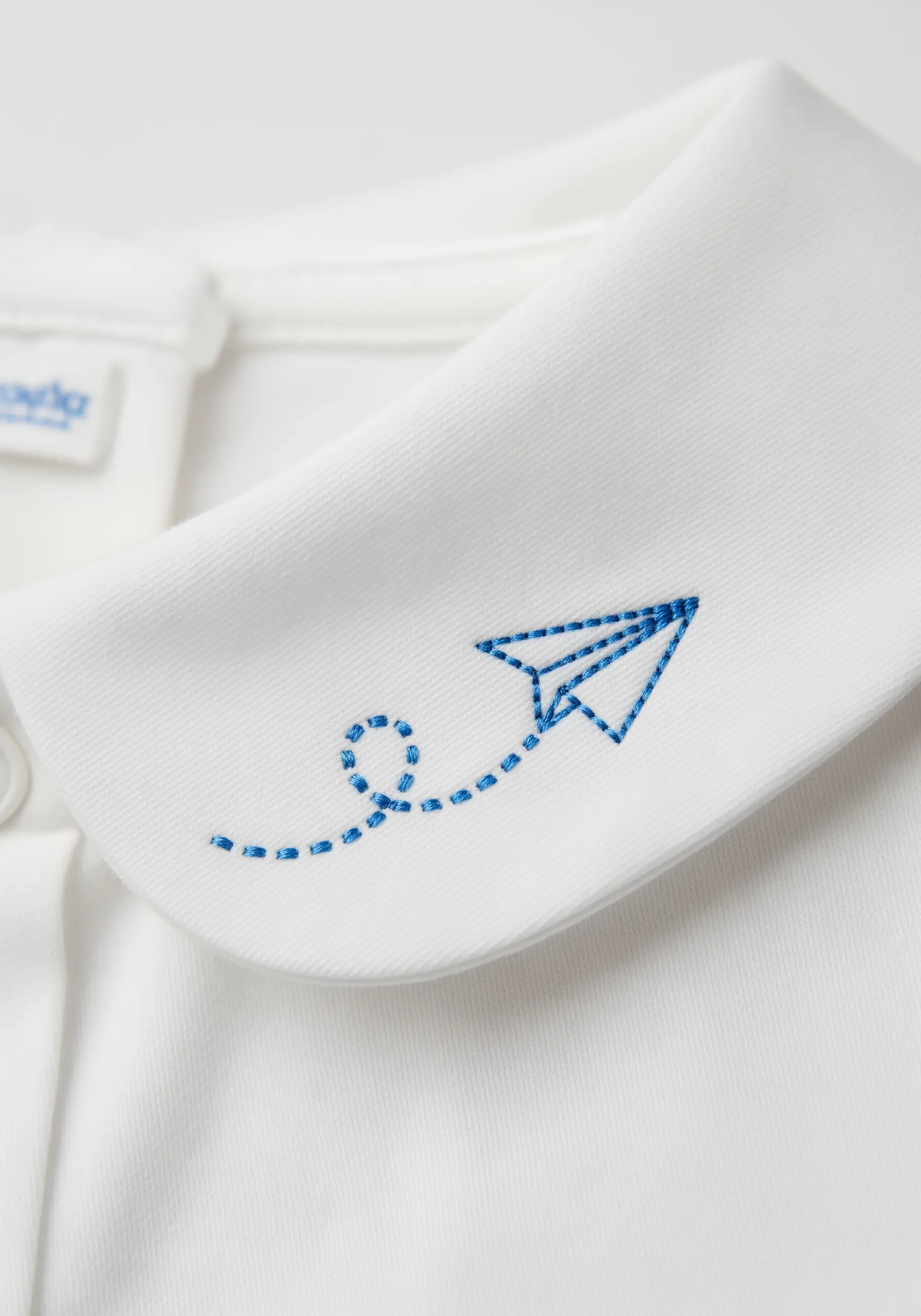 Blue embroidered paper airplane with a running stitch flight path on a white collar.