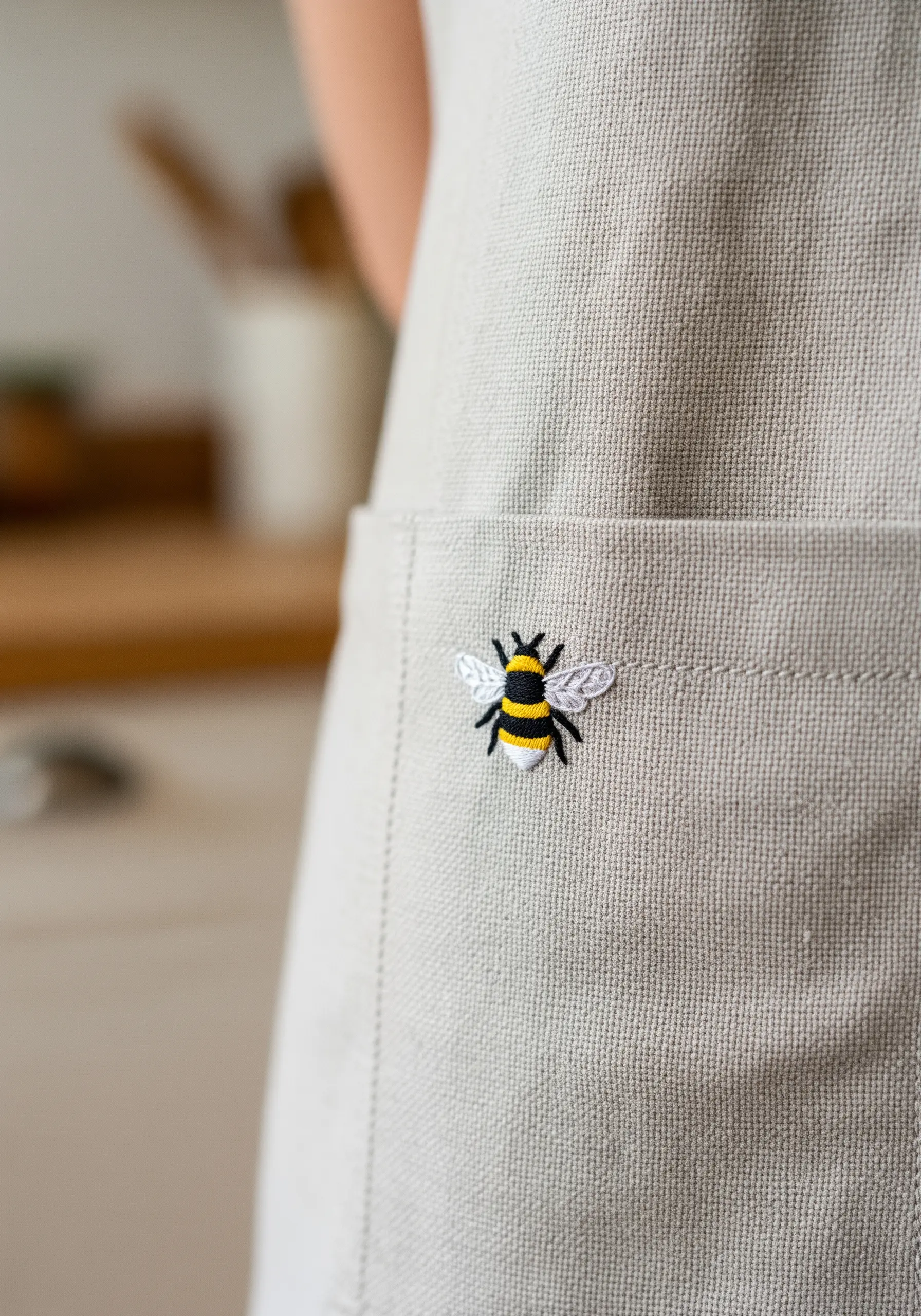 A tiny, detailed bee embroidered on the pocket of a neutral-colored apron.