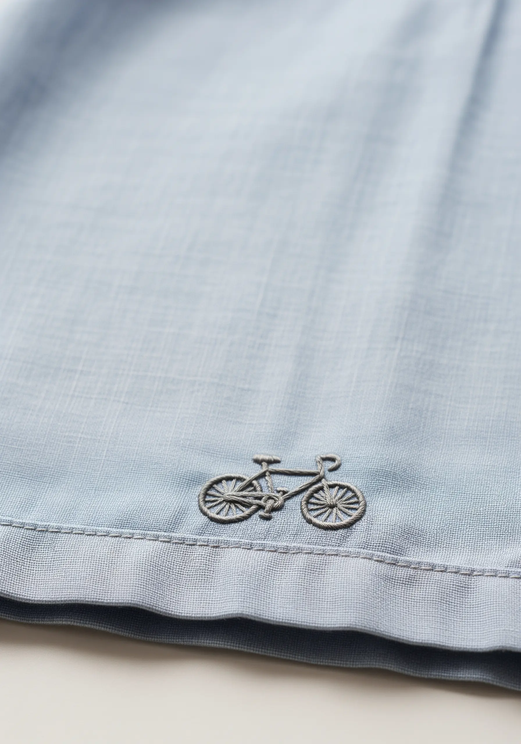 A tiny, detailed bicycle embroidered in grey thread on the hem of a light blue shirt.