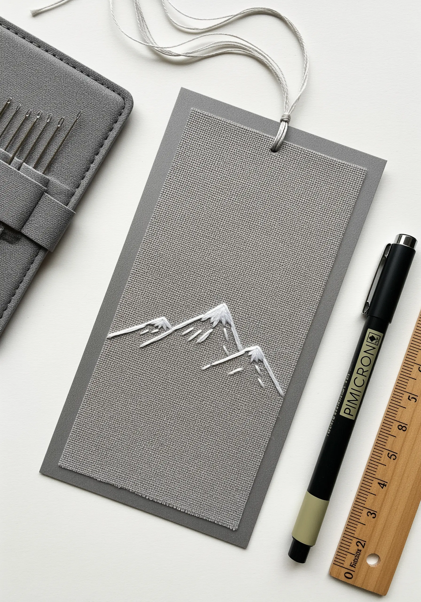 Minimalist white thread mountain range embroidered on grey Aida cloth, mounted on a bookmark.