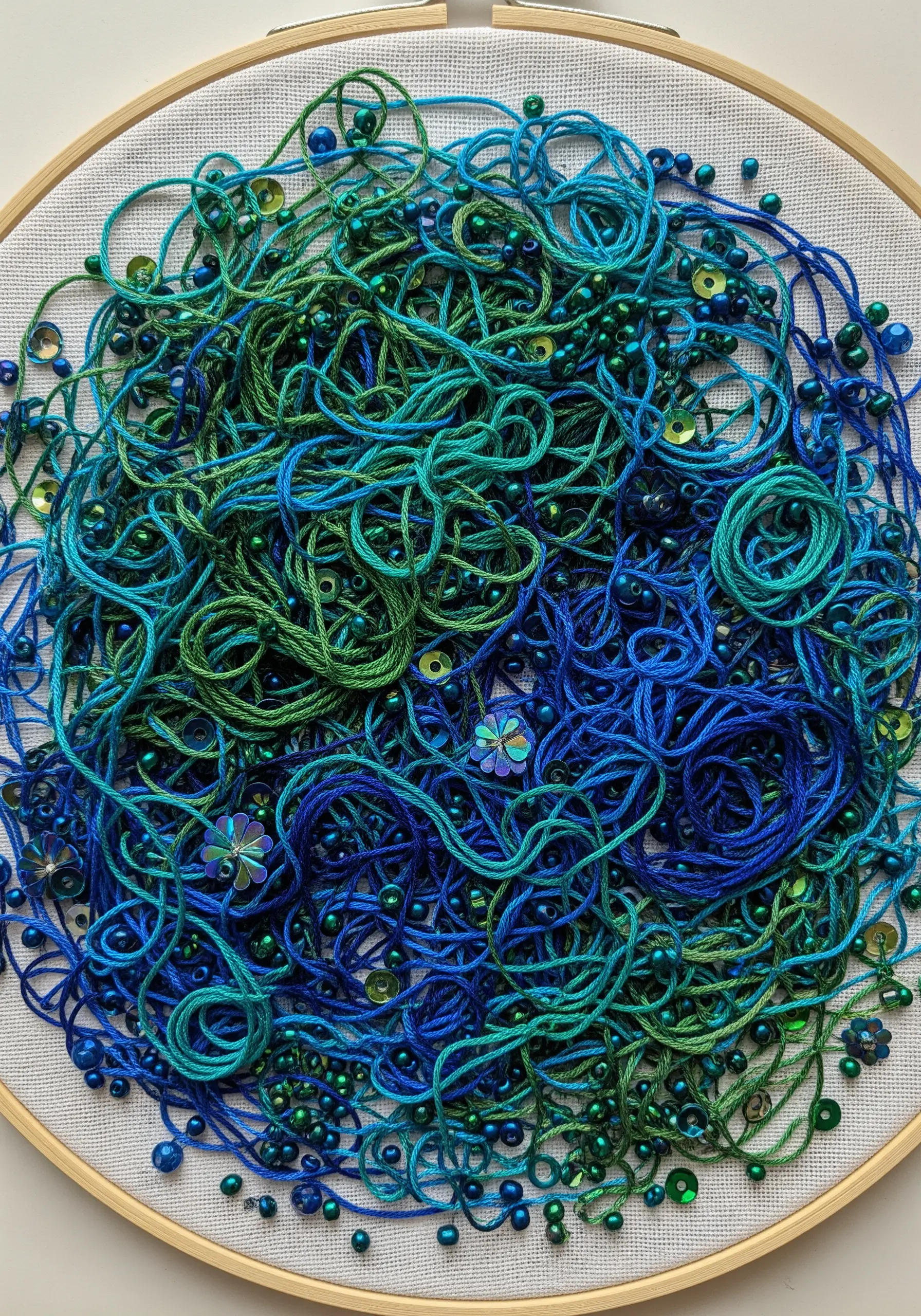 A hoop filled with a chaotic mix of green and blue threads, interspersed with shimmering beads and sequins