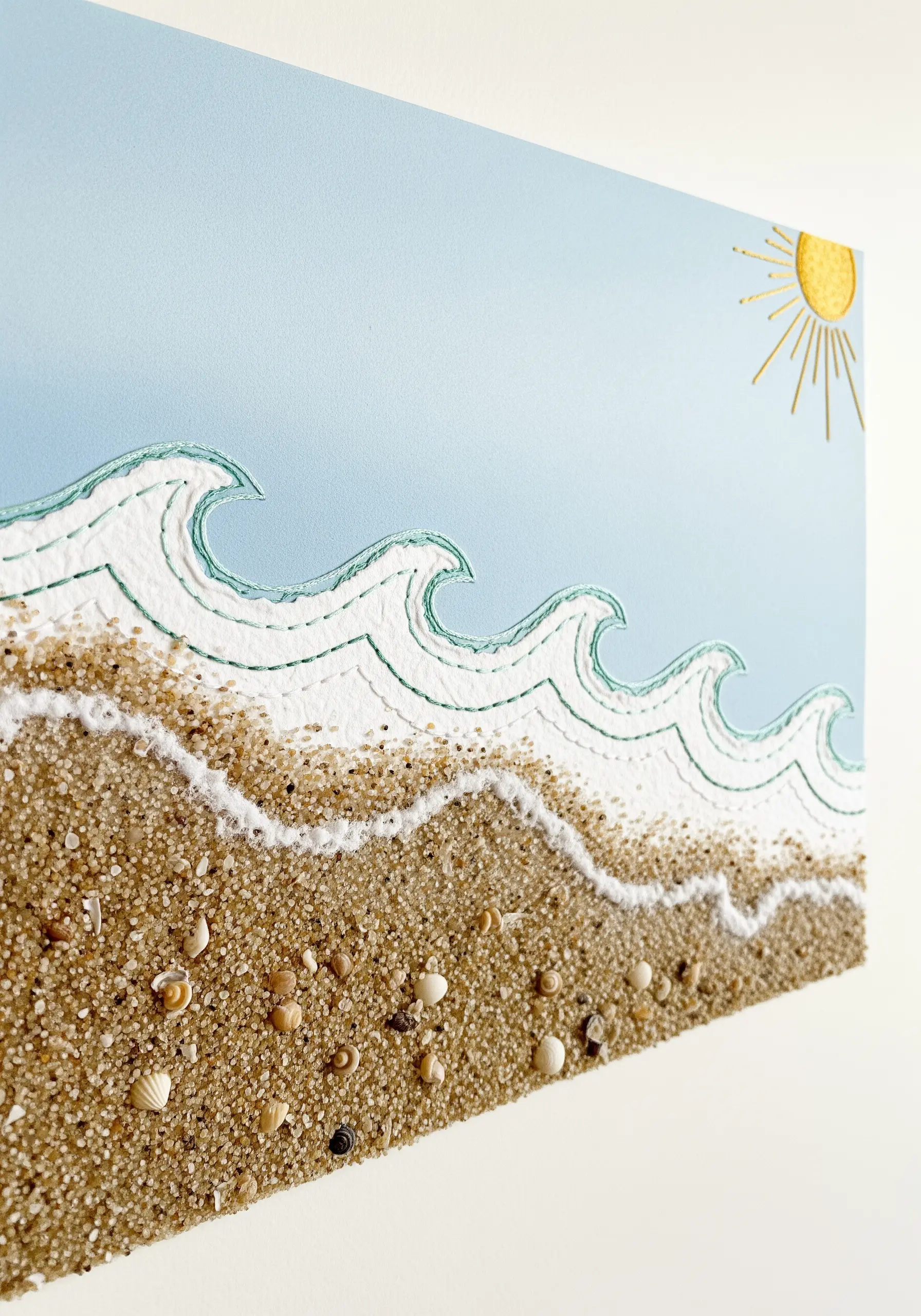 Embroidered waves on paper with real sand and shells creating a 3D beach.