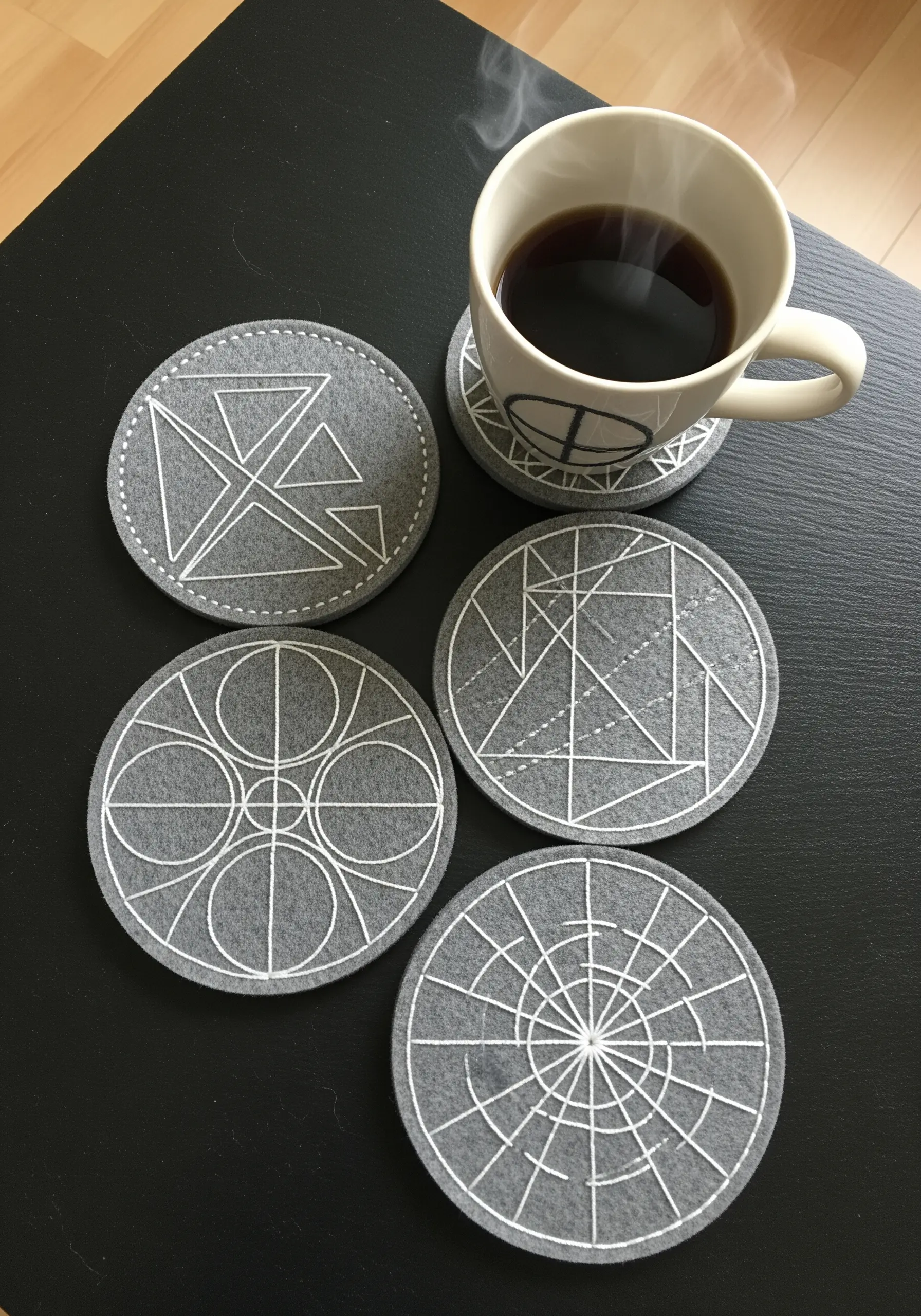 A set of gray felt coasters with modern geometric patterns stitched in white thread.
