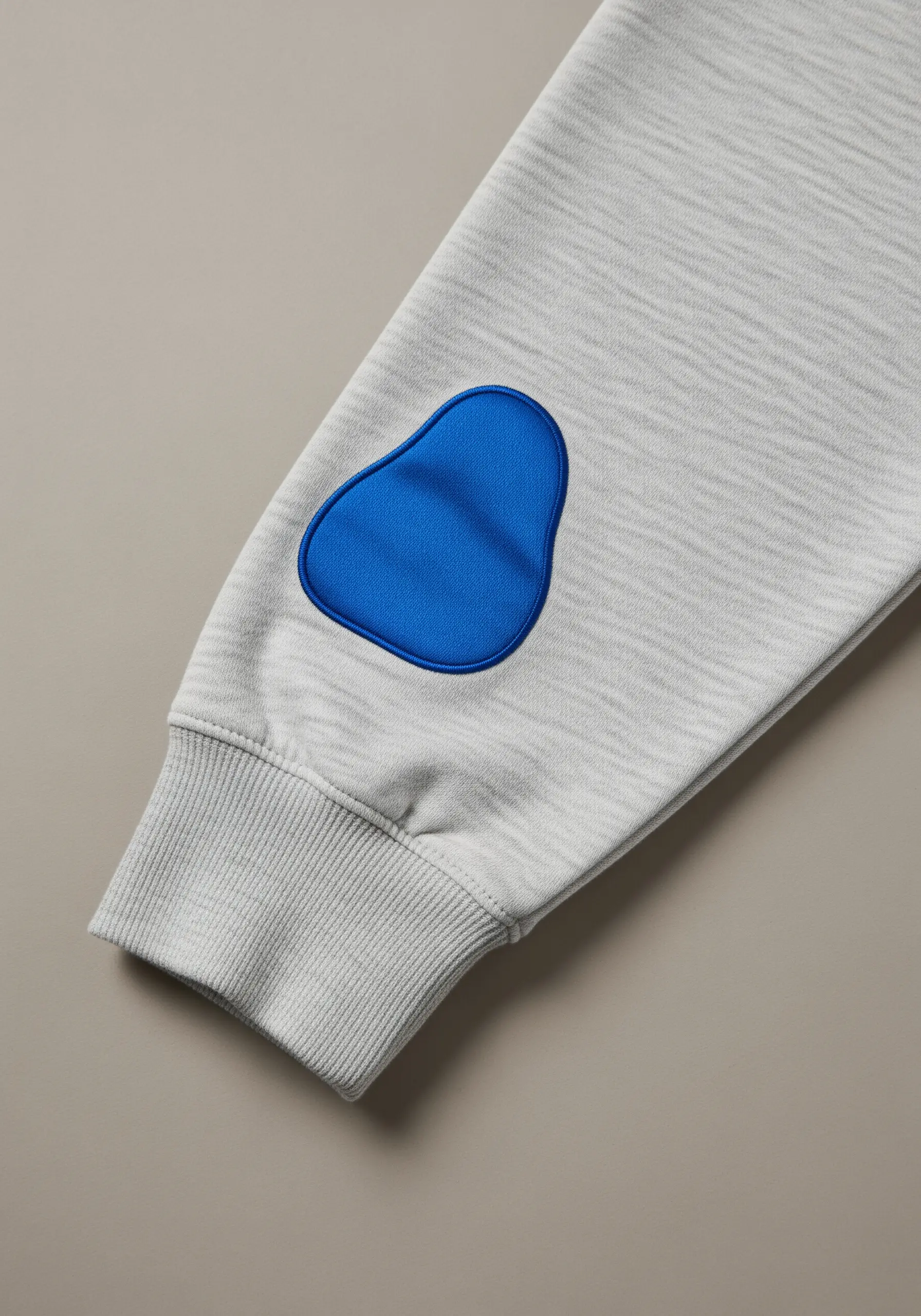 A modern, abstract-shaped elbow patch in bright blue satin stitch on a grey sweatshirt.