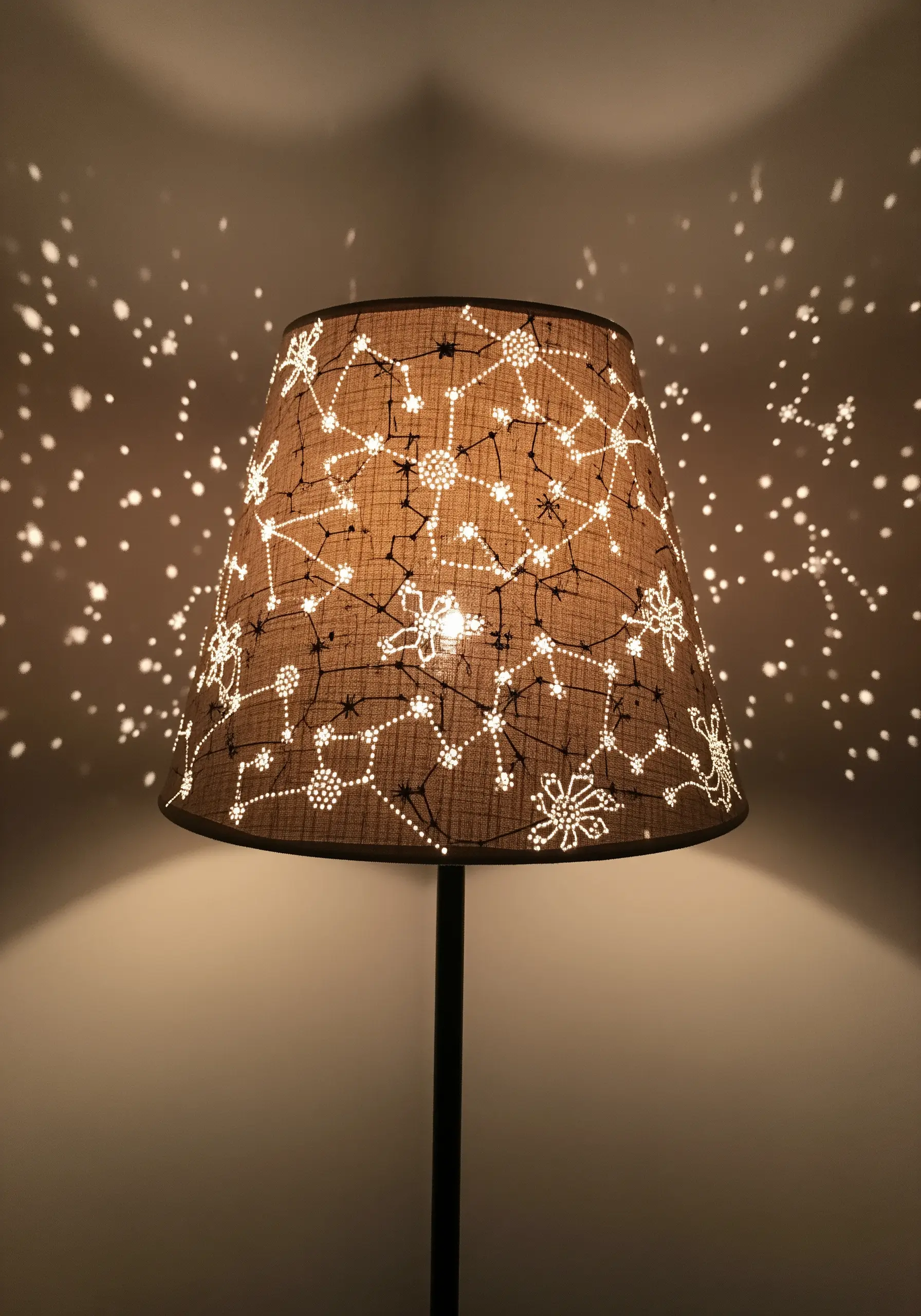 A lampshade with an embroidered constellation pattern, perforated to project points of light onto the wall.