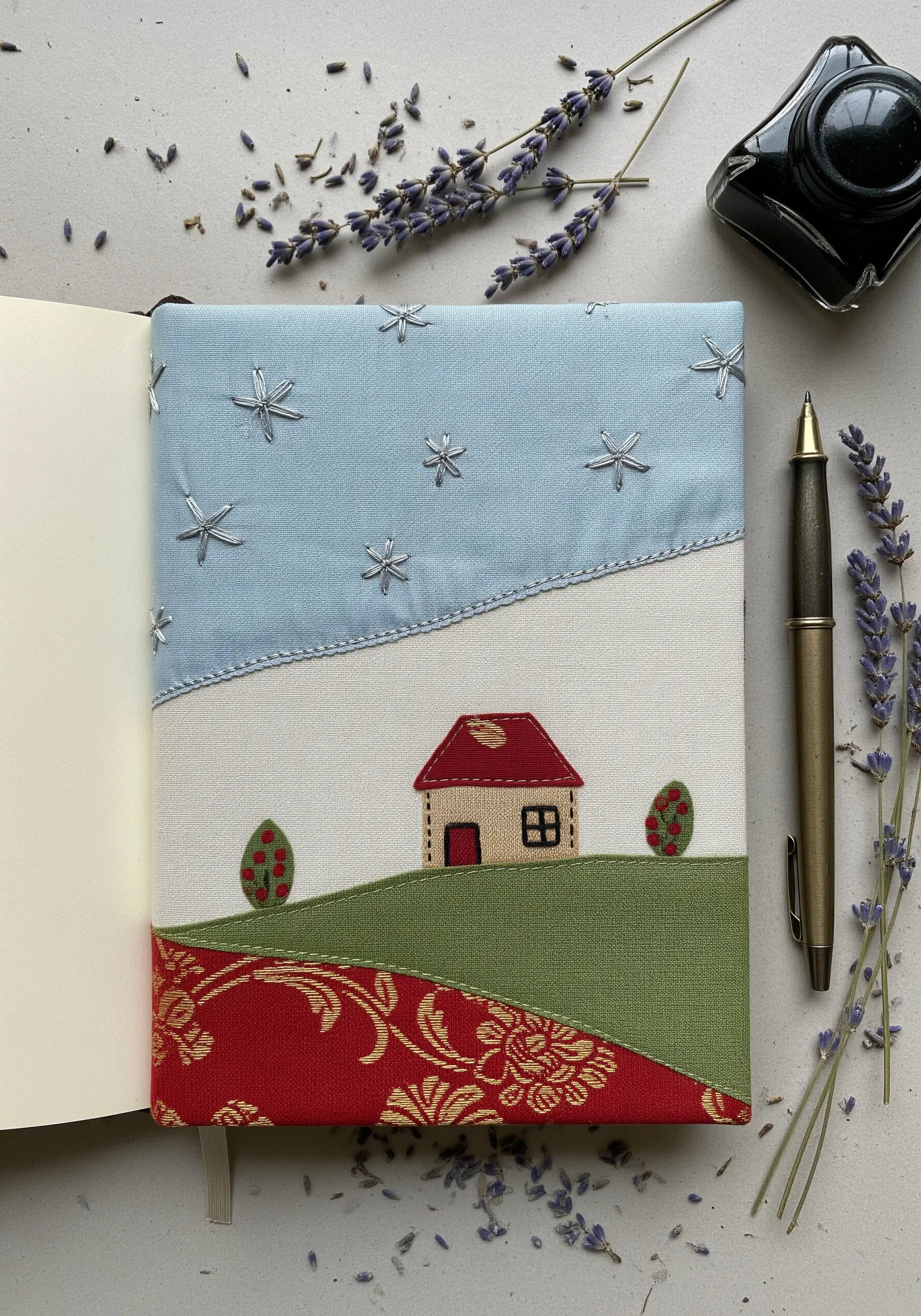 Embroidered journal cover with a fabric collage landscape of a house on a hill.