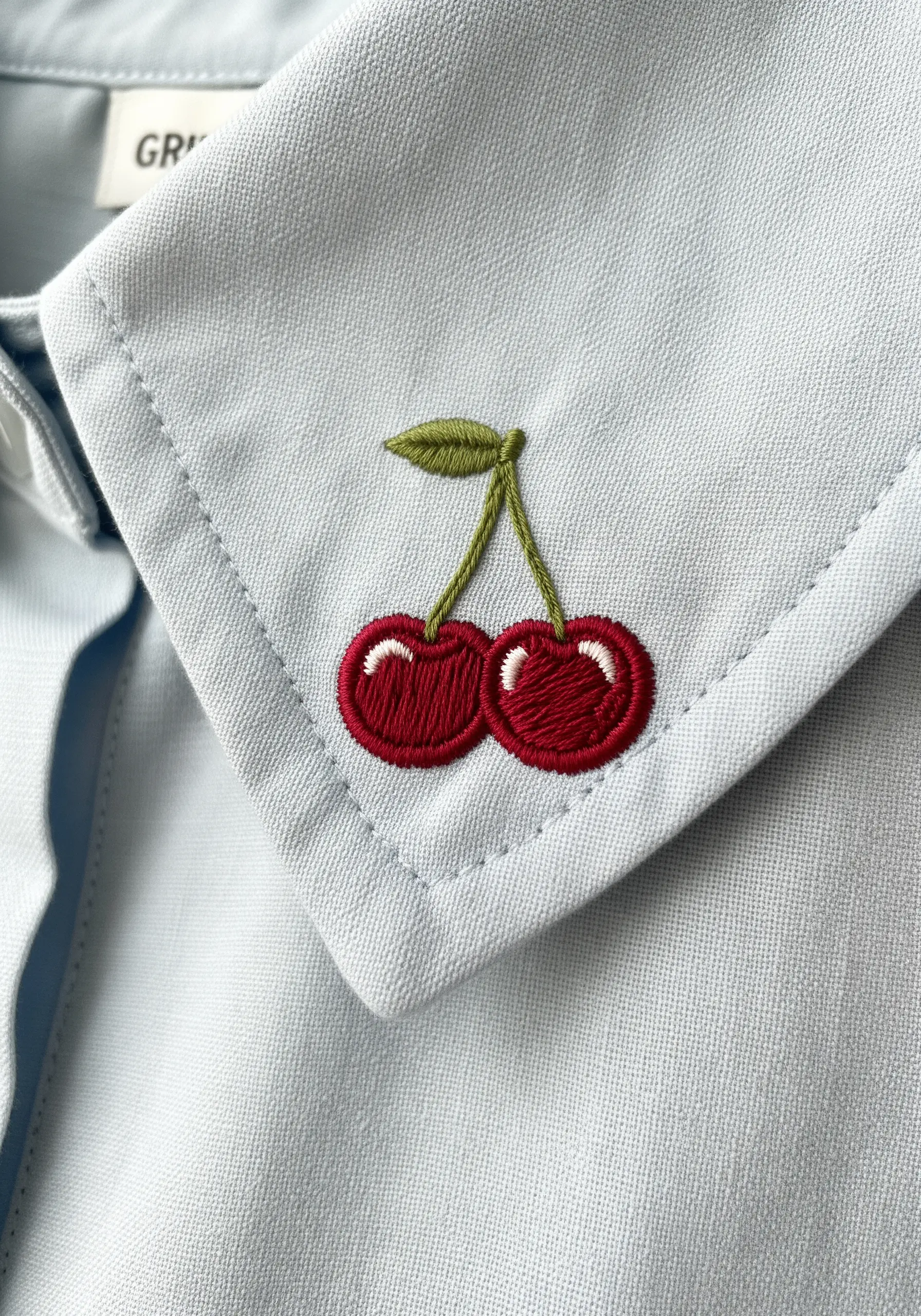 A pair of embroidered cherries with a clean white highlight on a light blue shirt collar.