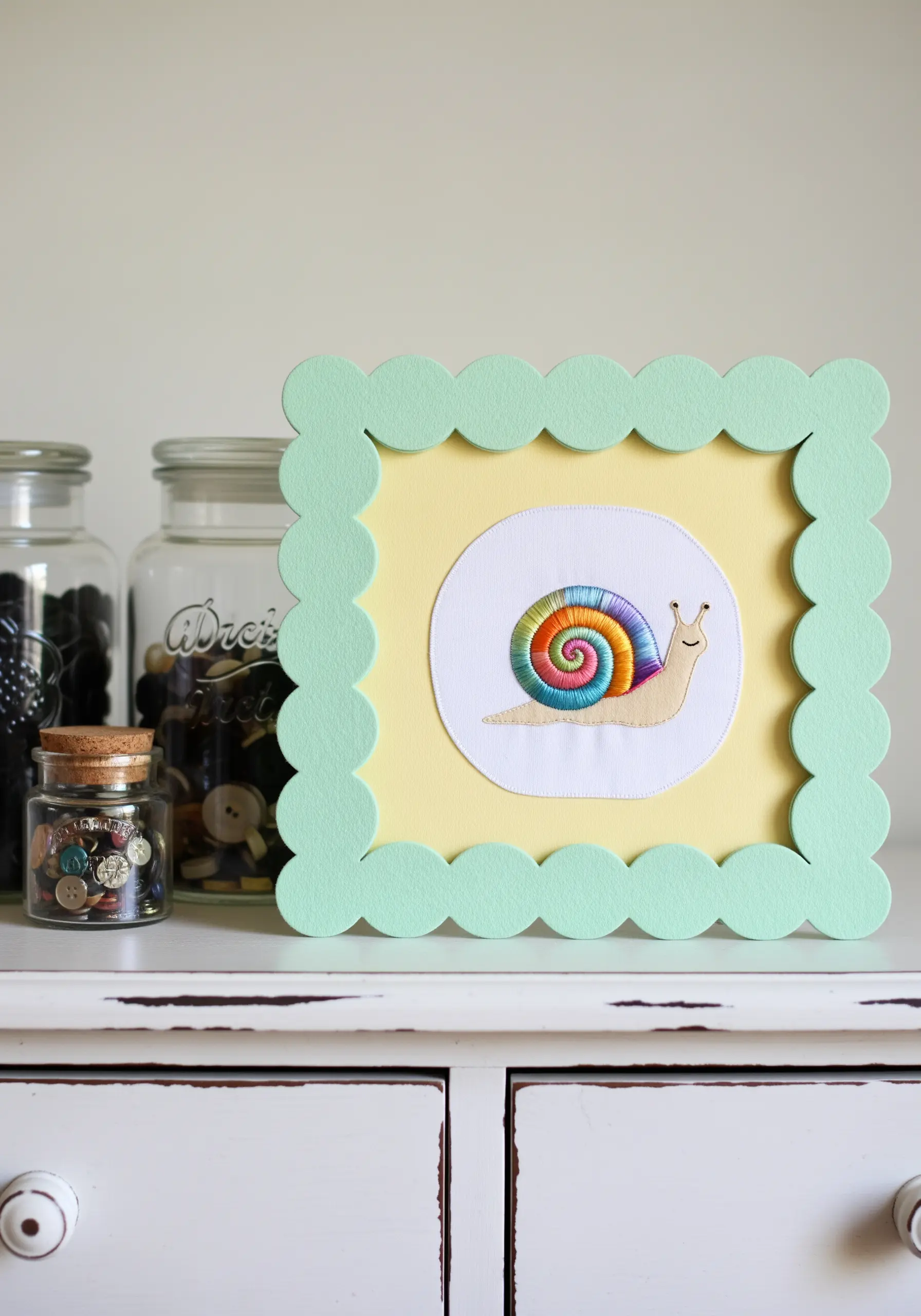 An embroidered snail with a rainbow shell, matted on yellow paper with a mint green scalloped frame.