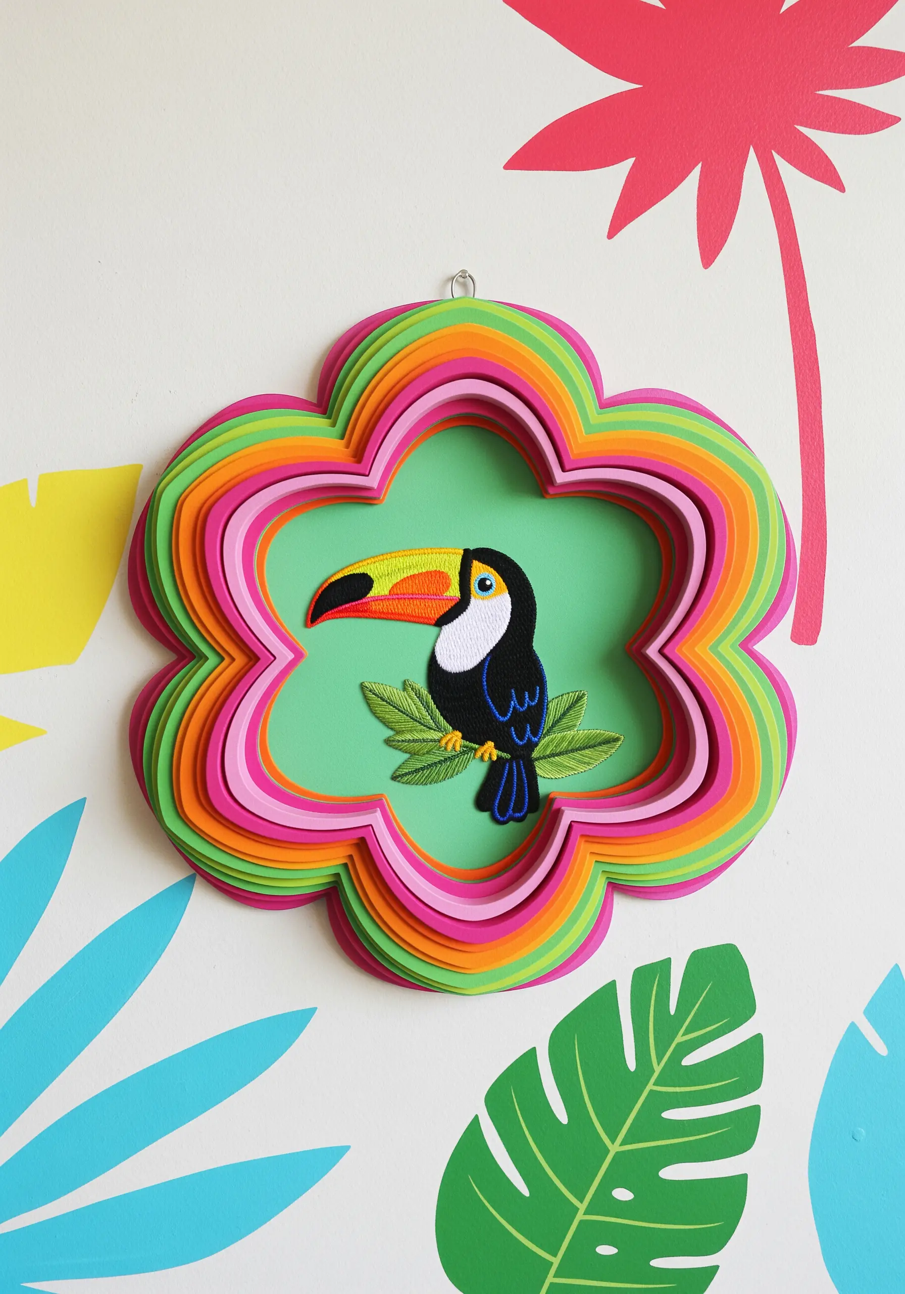 An embroidered toucan inside a vibrant, layered paper frame shaped like a flower.