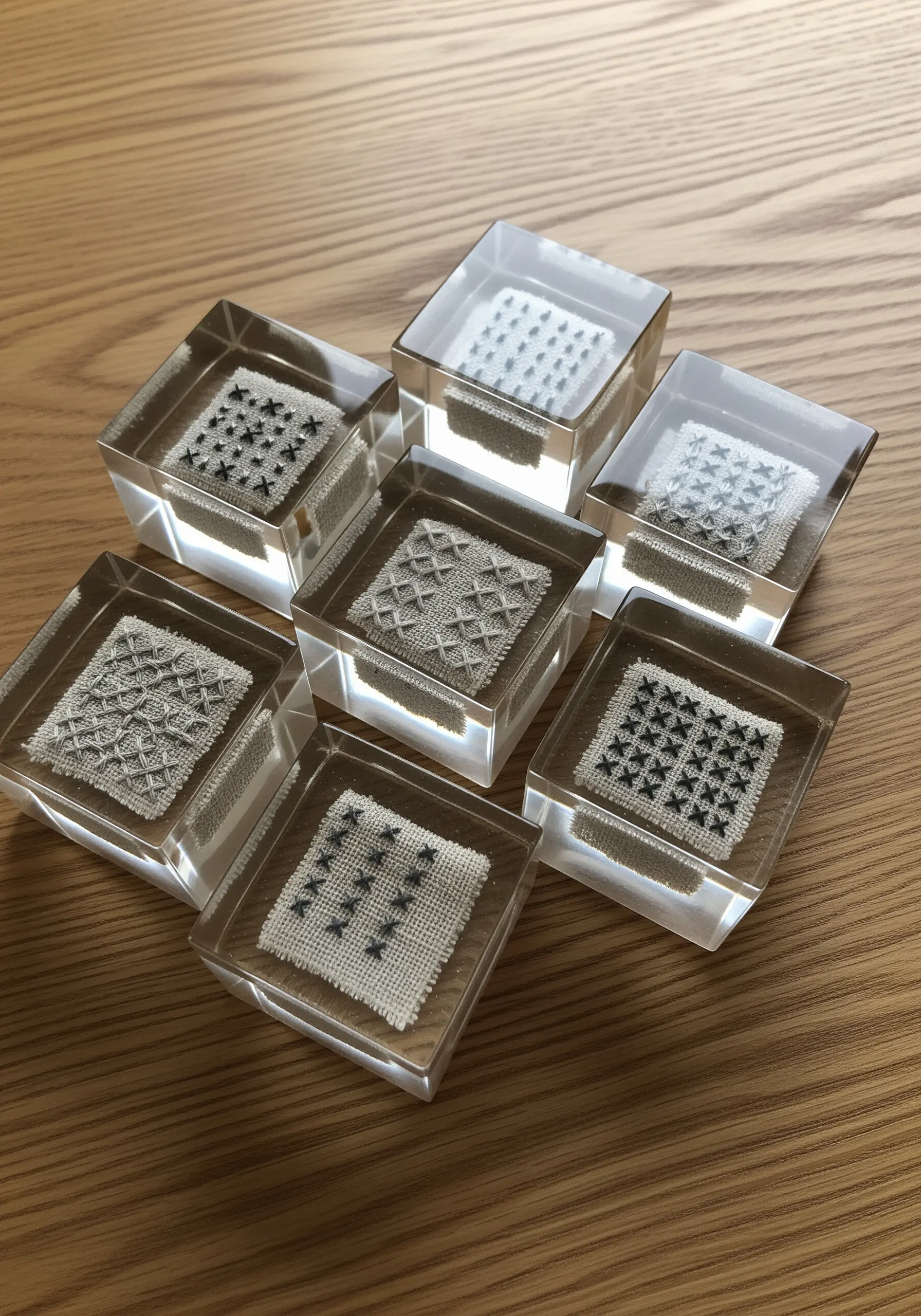 Small, monochrome cross-stitch samples preserved in clear resin cubes on a wooden surface.