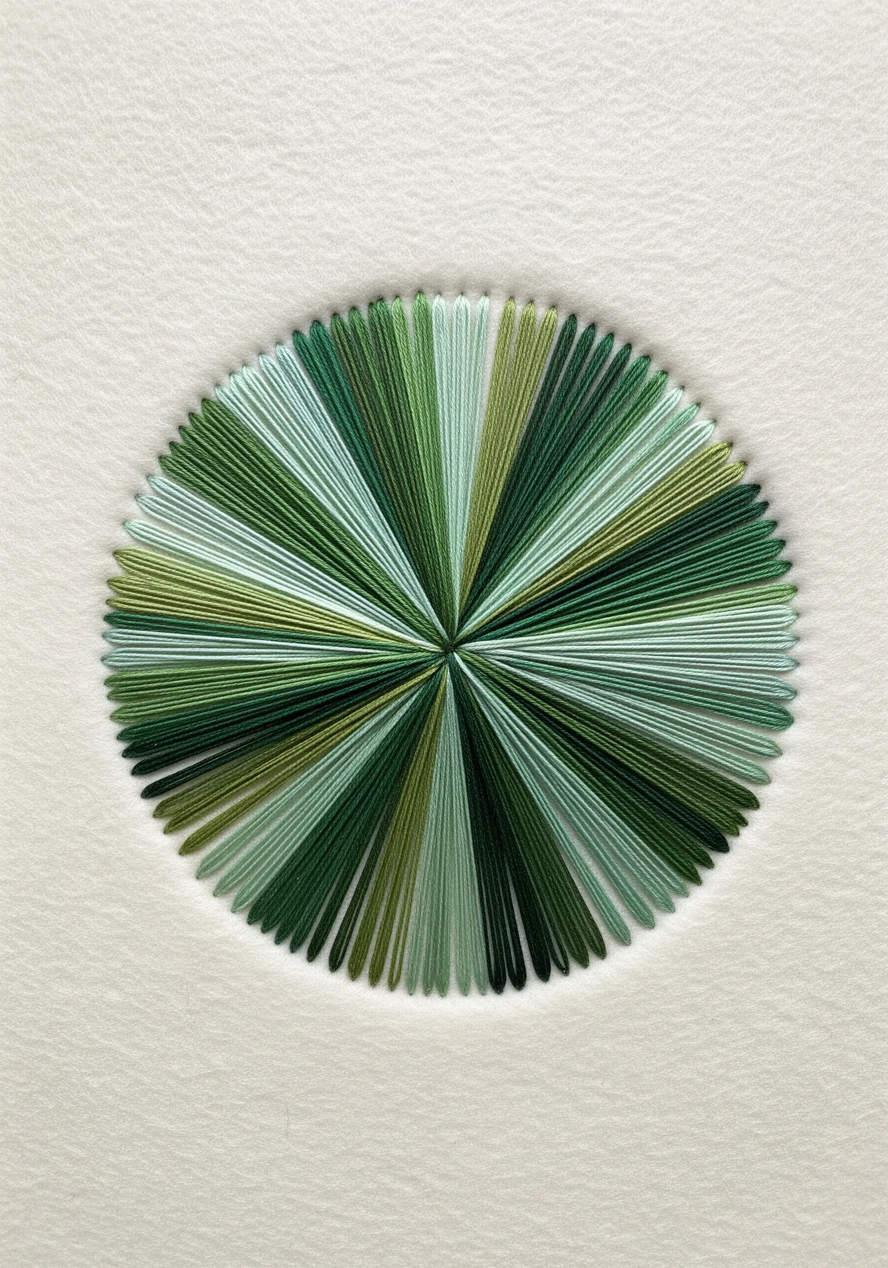 A perfect circle of radiating straight stitches in various shades of green on a white background