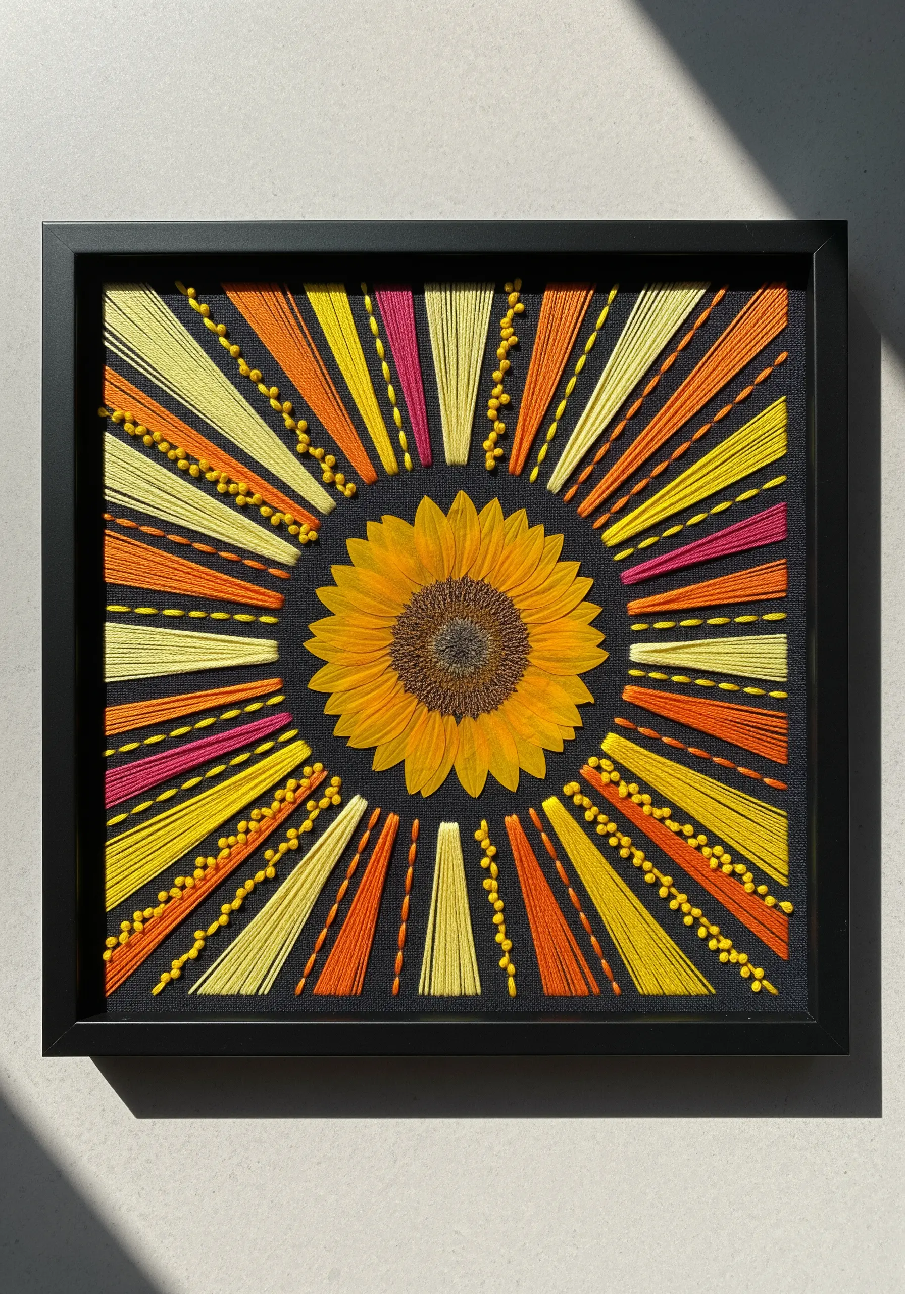 A pressed sunflower at the center of a radiating sunburst of yellow, orange, and pink embroidery.