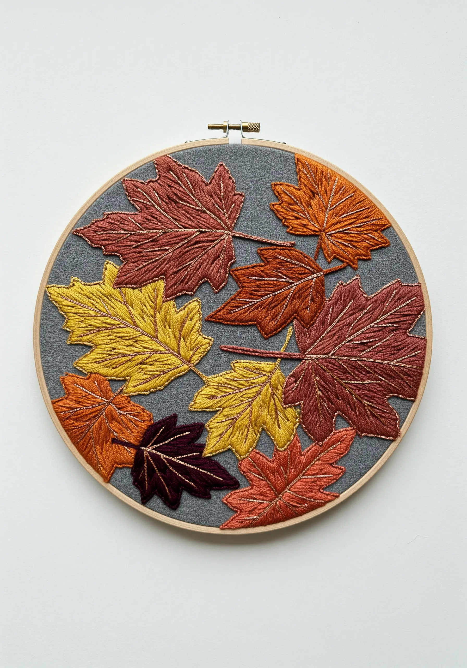 Embroidery hoop with satin-stitched autumn leaves in red, orange, and yellow on grey fabric.