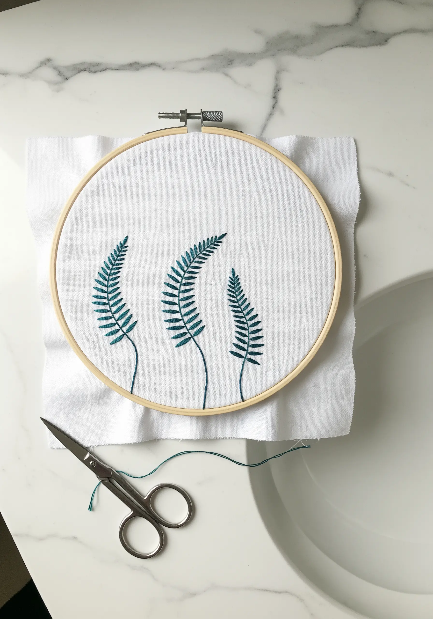Three minimalist fern fronds in teal thread on white fabric in a hoop.