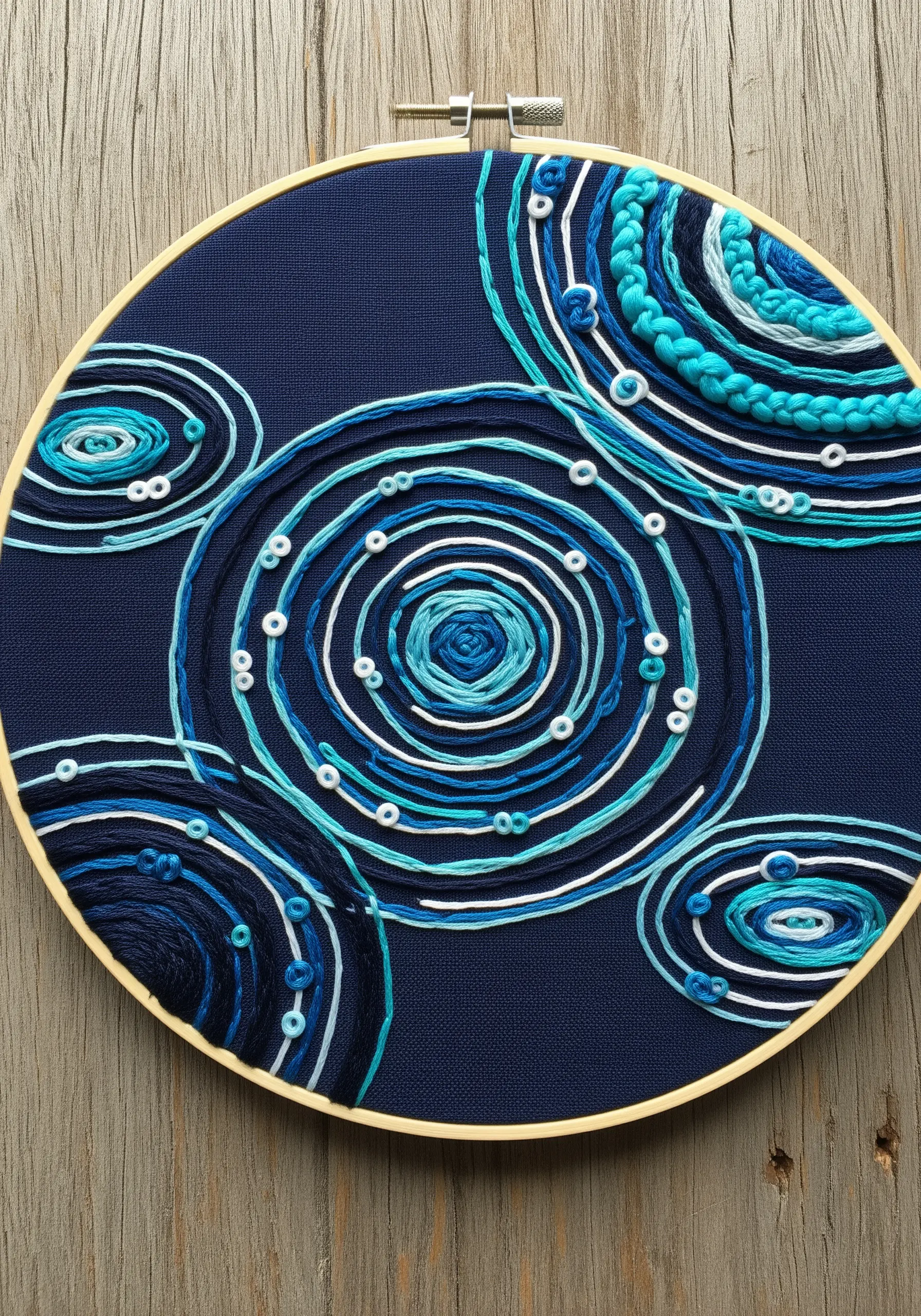 Abstract hoop art with concentric circles in blue and turquoise threads on dark navy fabric.