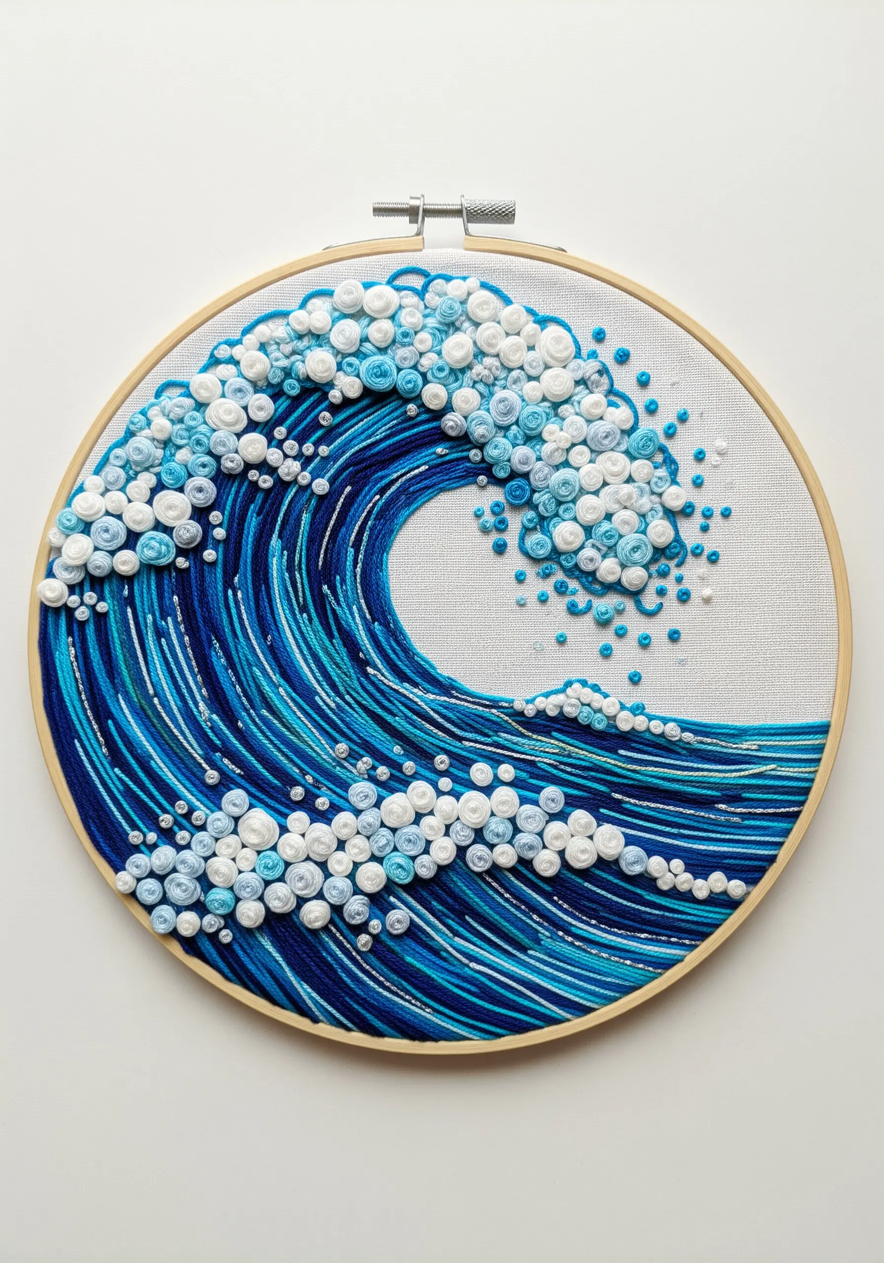 Embroidery hoop art of a large, cresting ocean wave made with blue threads and textured white knots.