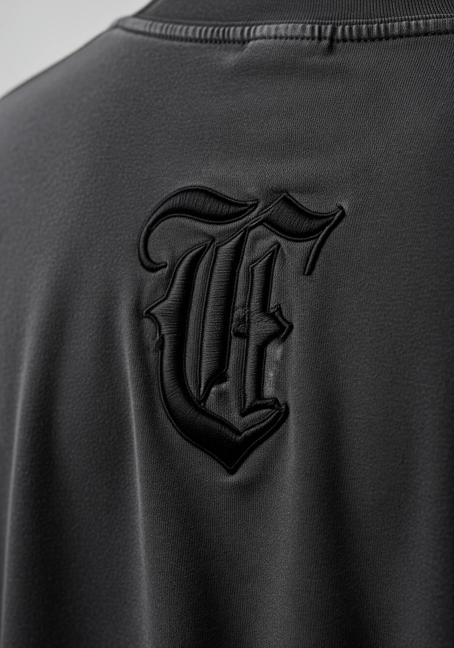 A black gothic letter 'E' embroidered on a black shirt, creating a raised, tone-on-tone effect.