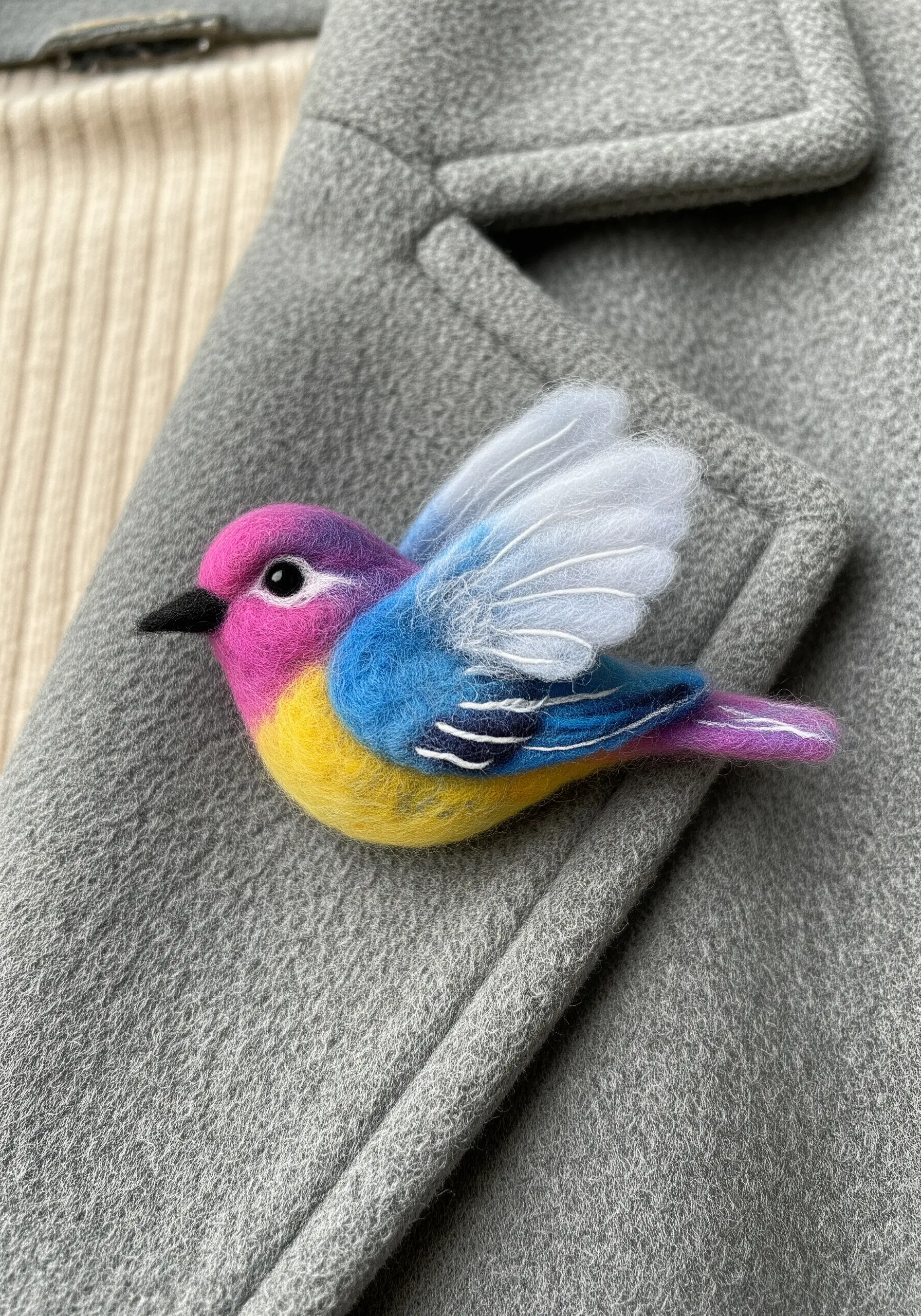 A colorful needle-felted bird brooch with delicate white wings, pinned to a grey coat lapel.