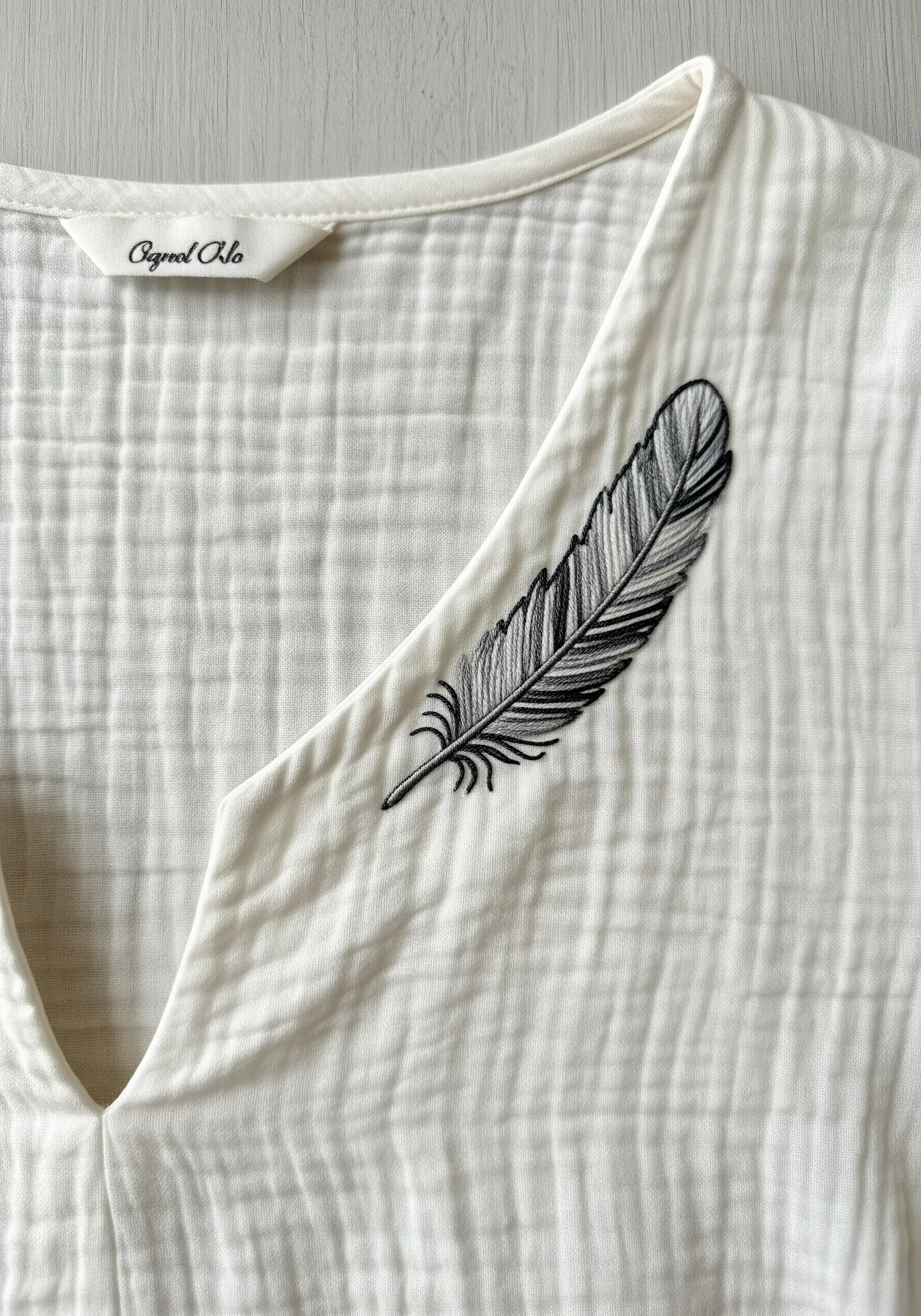 A detailed feather with black and gray shading embroidered on a white muslin shirt.