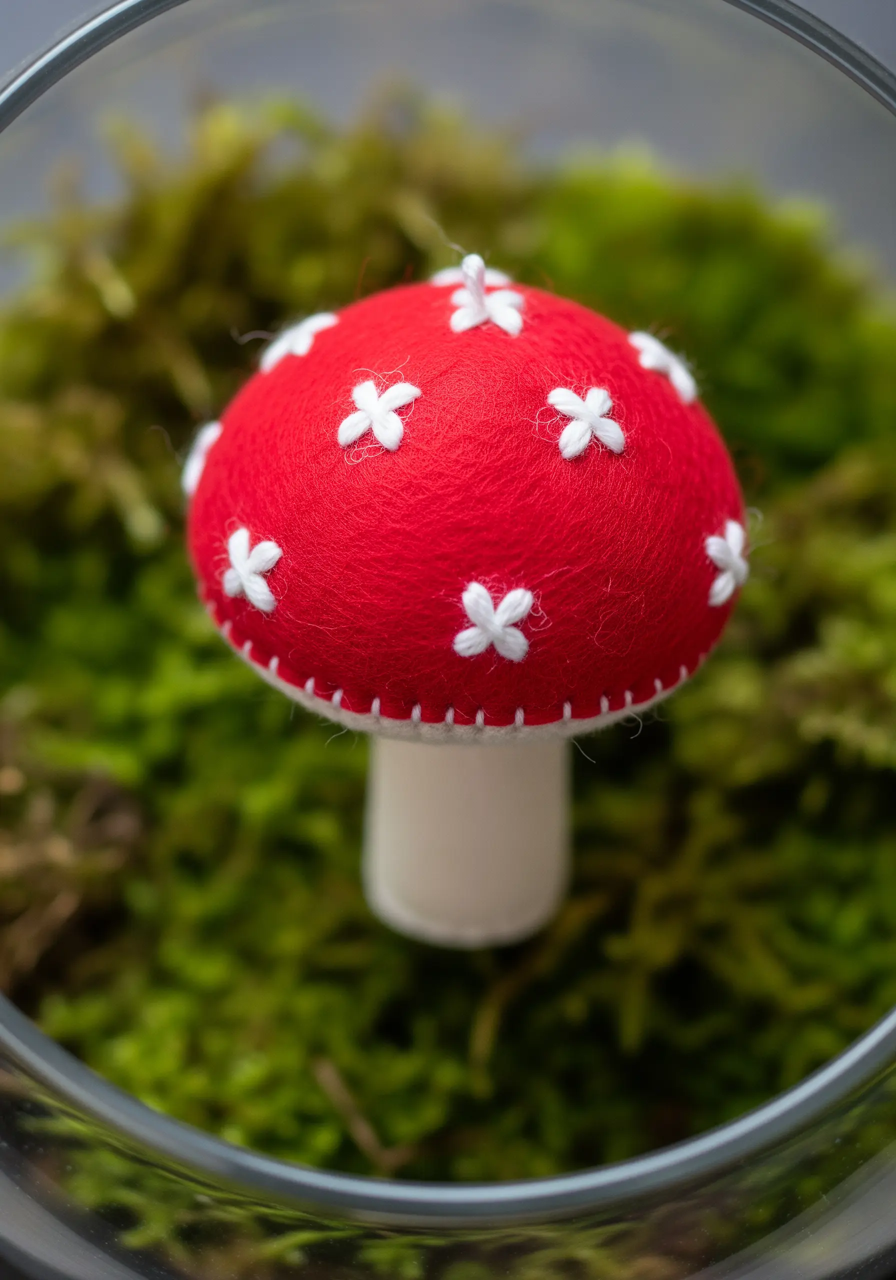 A small, 3D felt toadstool with a red cap and white cross-stitched spots, sitting on moss.