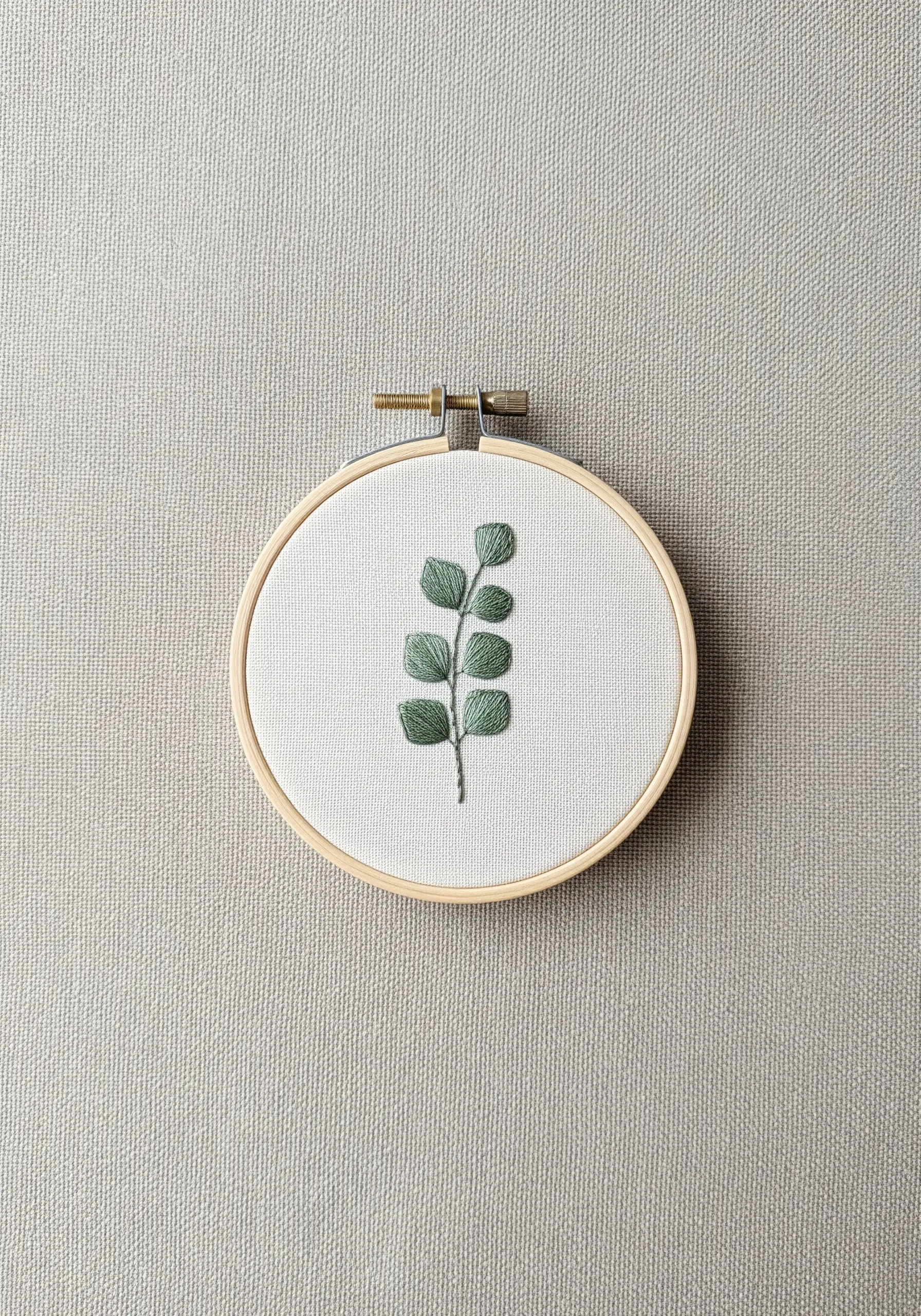 A single eucalyptus branch with satin-stitched leaves on light fabric in a hoop.