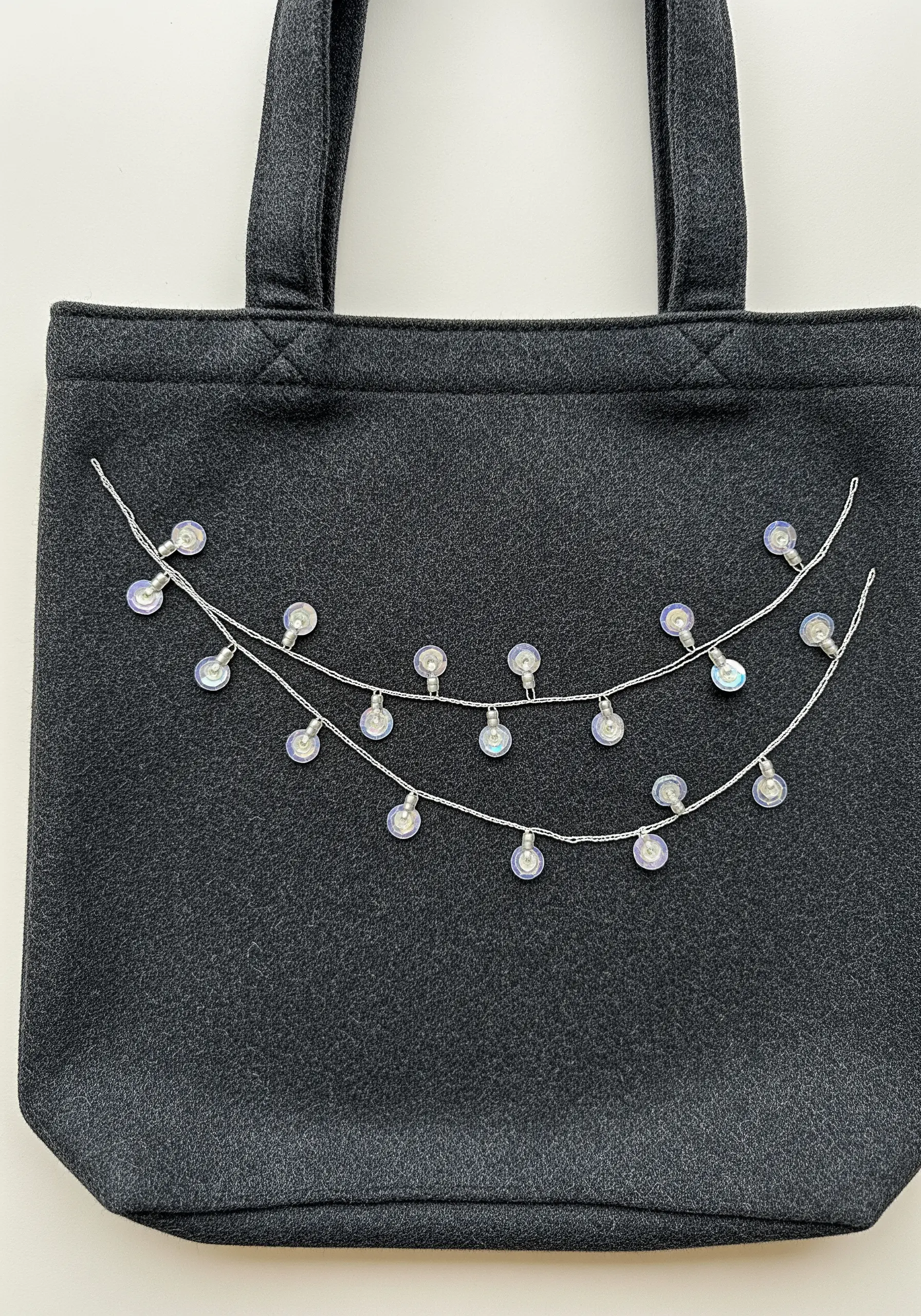 Minimalist holiday lights made with silver thread and iridescent sequins on a dark gray felt tote.