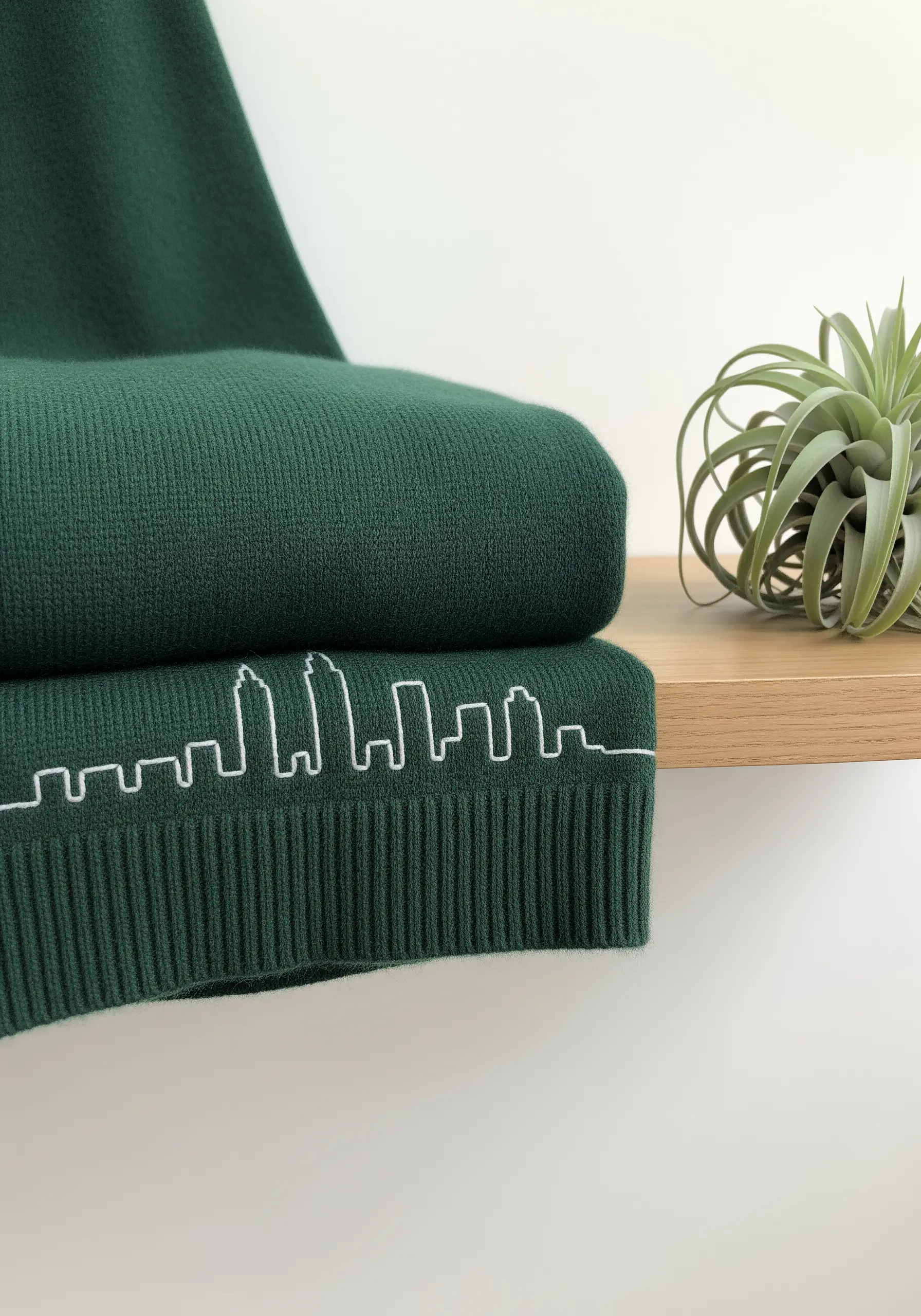 A white line-art city skyline embroidered on the ribbed hem of a dark green sweater.