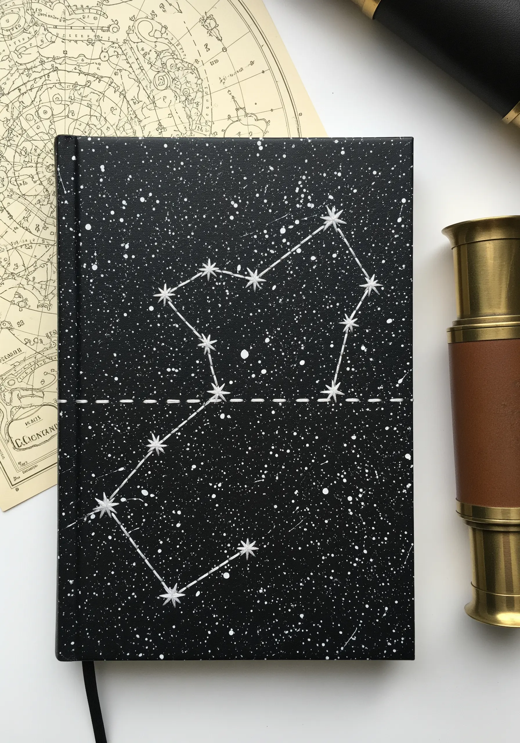 An embroidered star constellation in white thread on a black journal cover with white paint splatters.