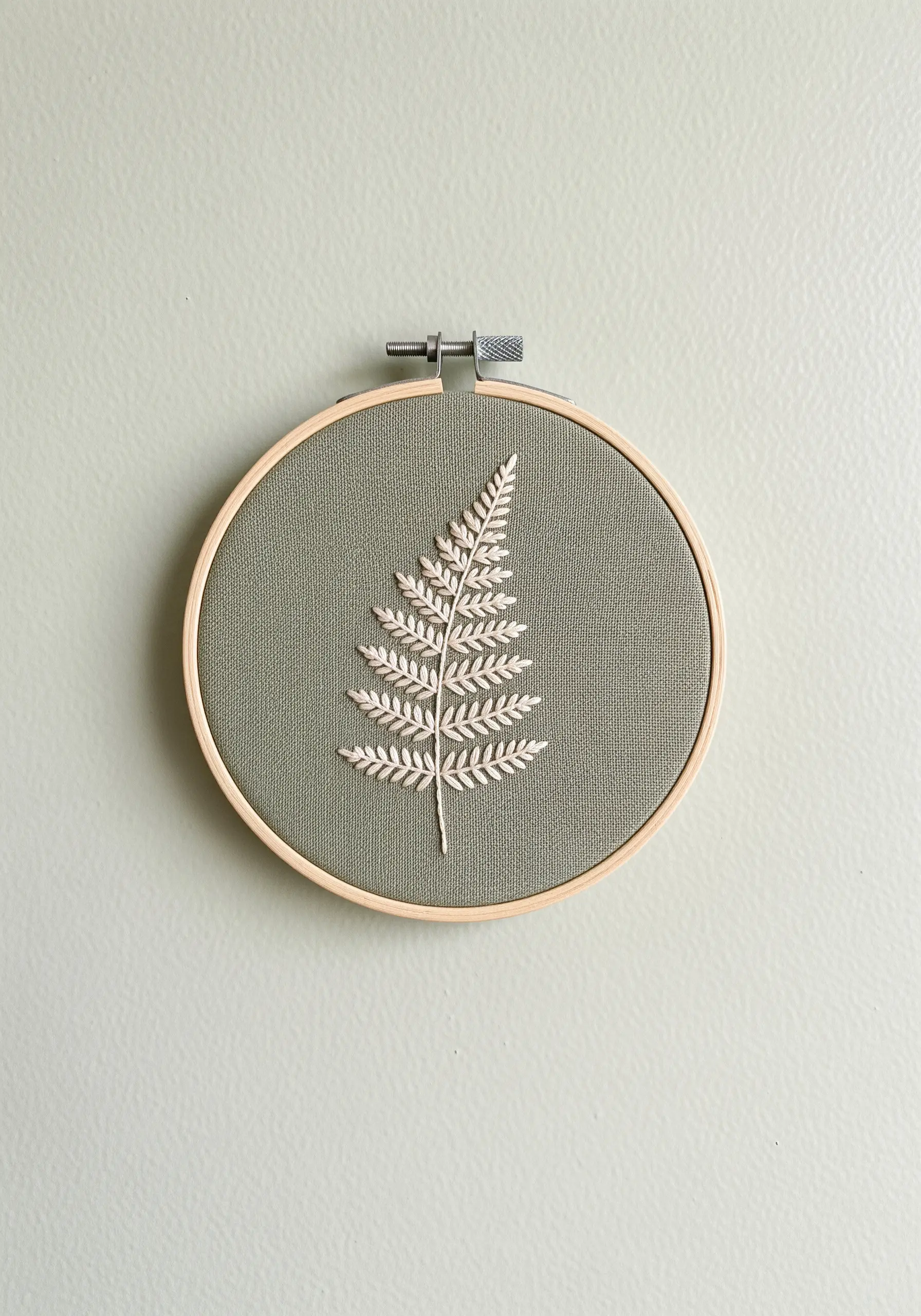 A delicate fern leaf embroidered in off-white thread on sage green fabric in a hoop.