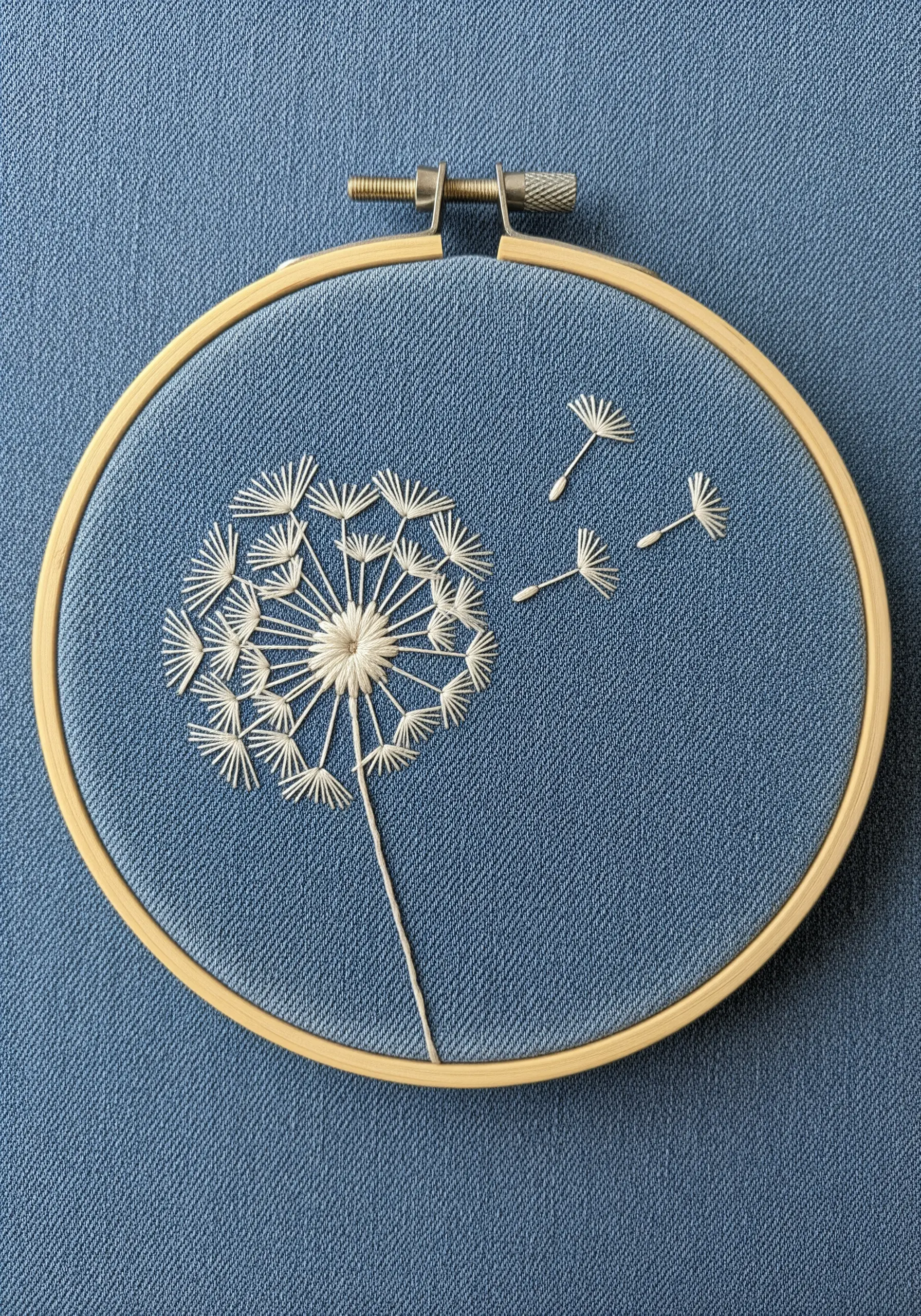 A delicate white dandelion with seeds blowing away, embroidered on blue fabric in a hoop.