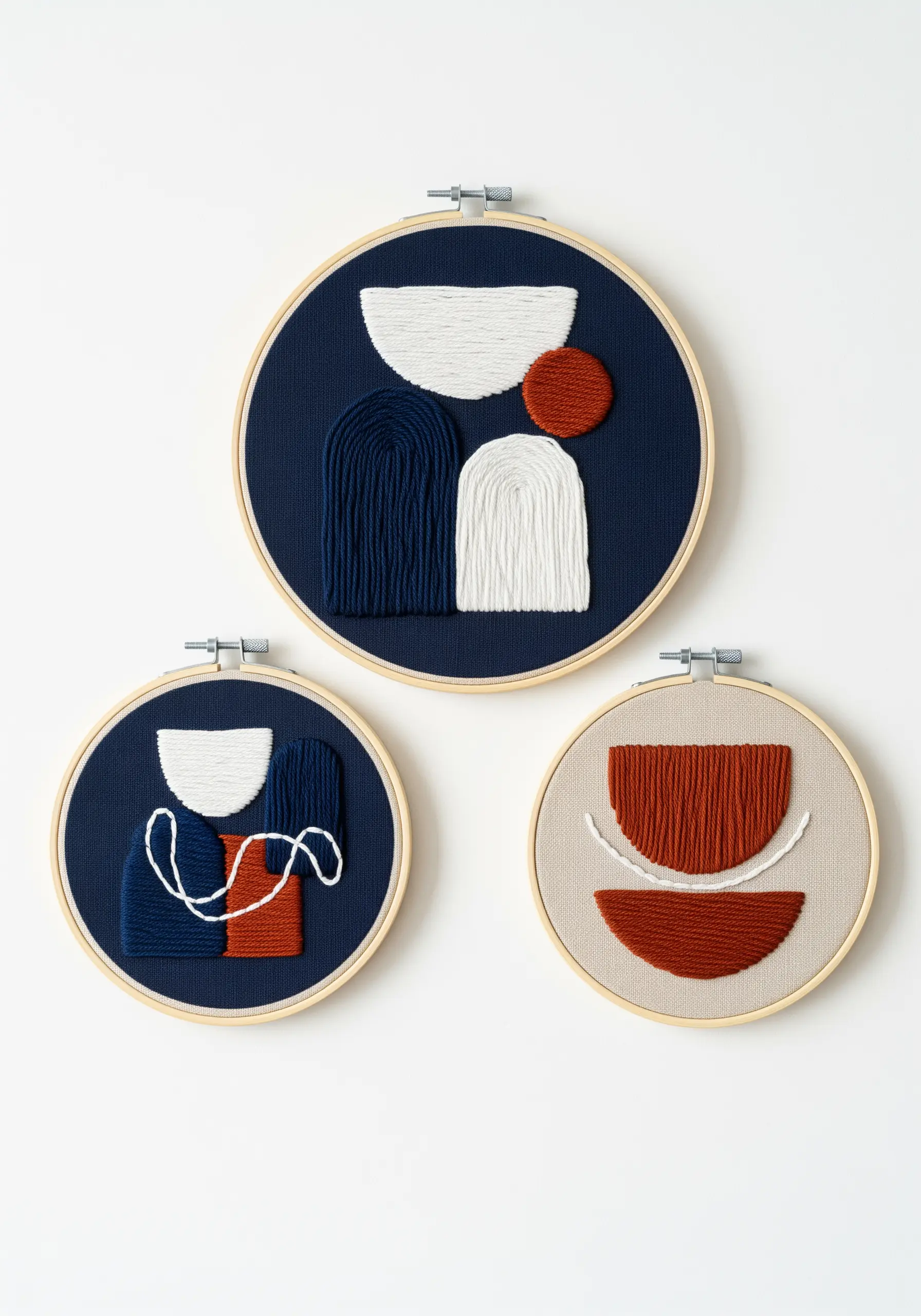 A trio of embroidery hoops with abstract shapes stitched in white, blue, and terracotta yarn.