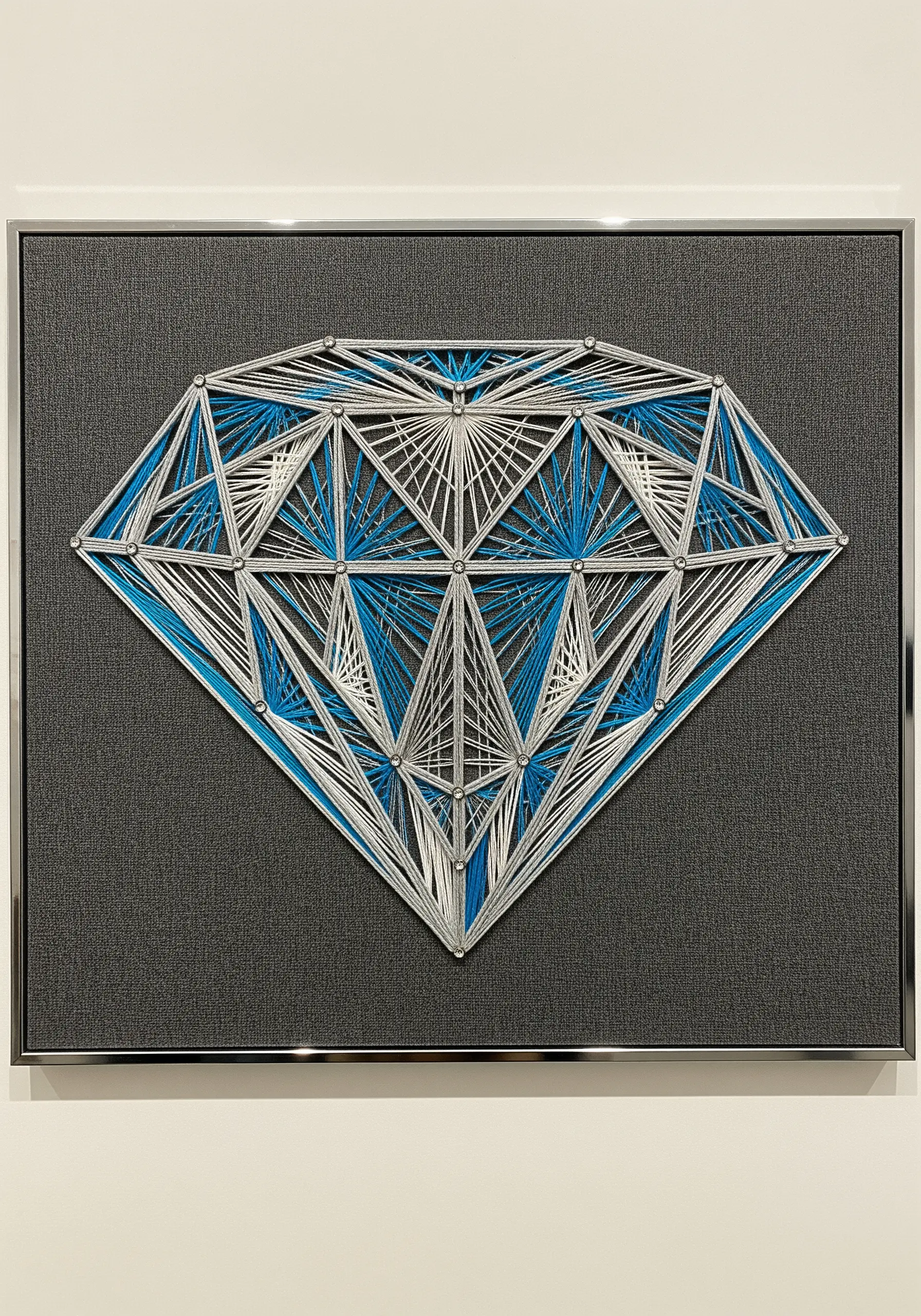 A geometric diamond embroidered with shiny white and blue thread on a gray textured background.