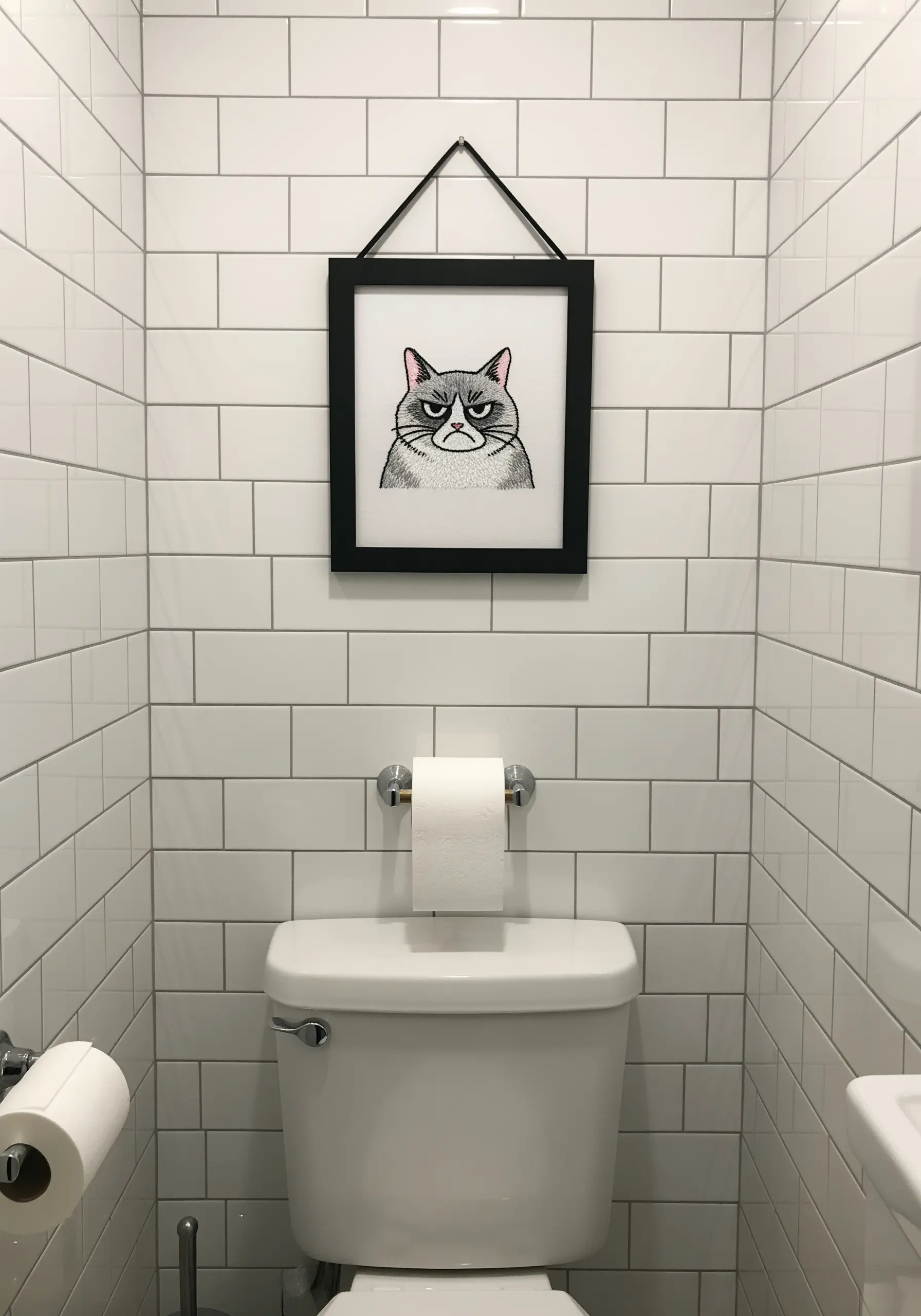 Embroidered grumpy cat portrait with a distinct frown, framed in a bathroom.