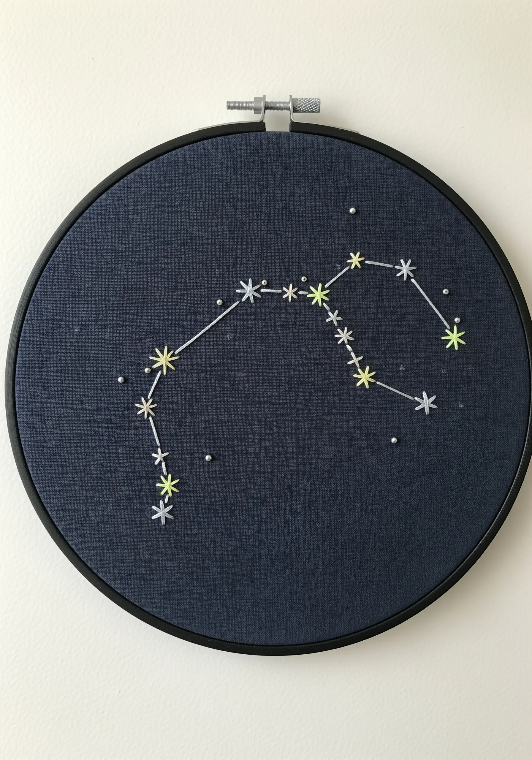 An embroidery hoop with a minimalist constellation stitched in light thread on dark blue fabric.