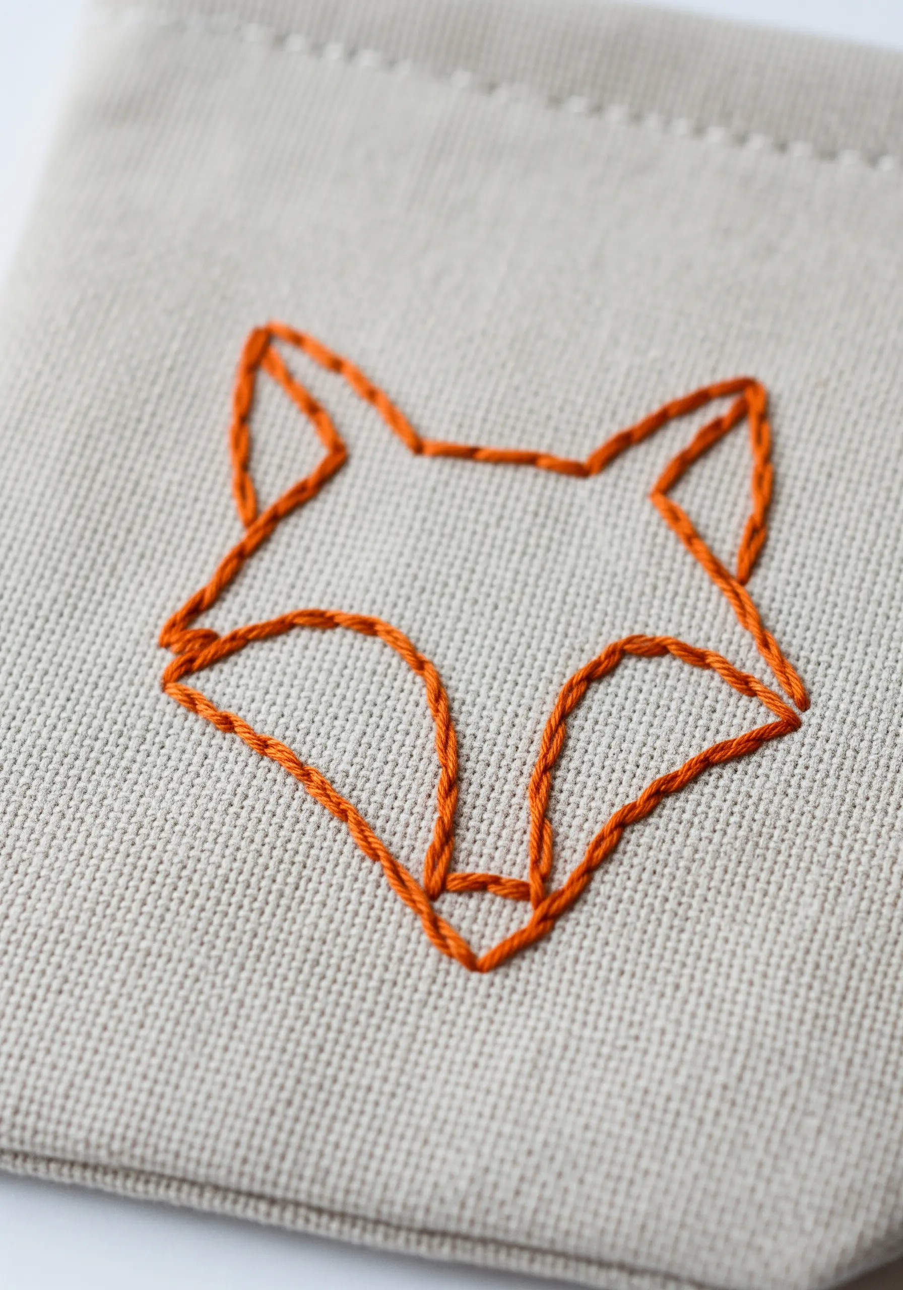 A minimalist geometric fox head outlined in orange thread on canvas.