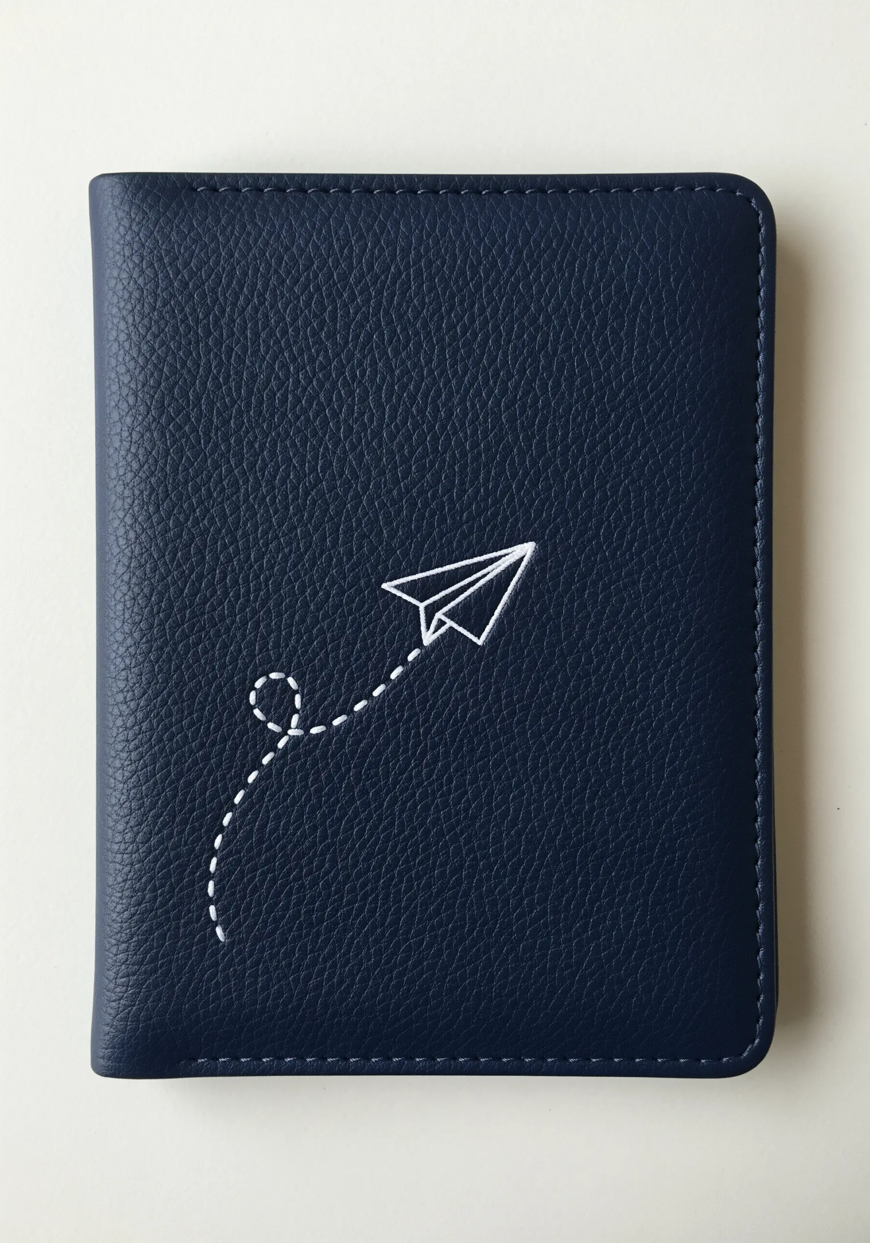 A paper airplane with a dotted flight path embroidered in white on a navy passport holder.