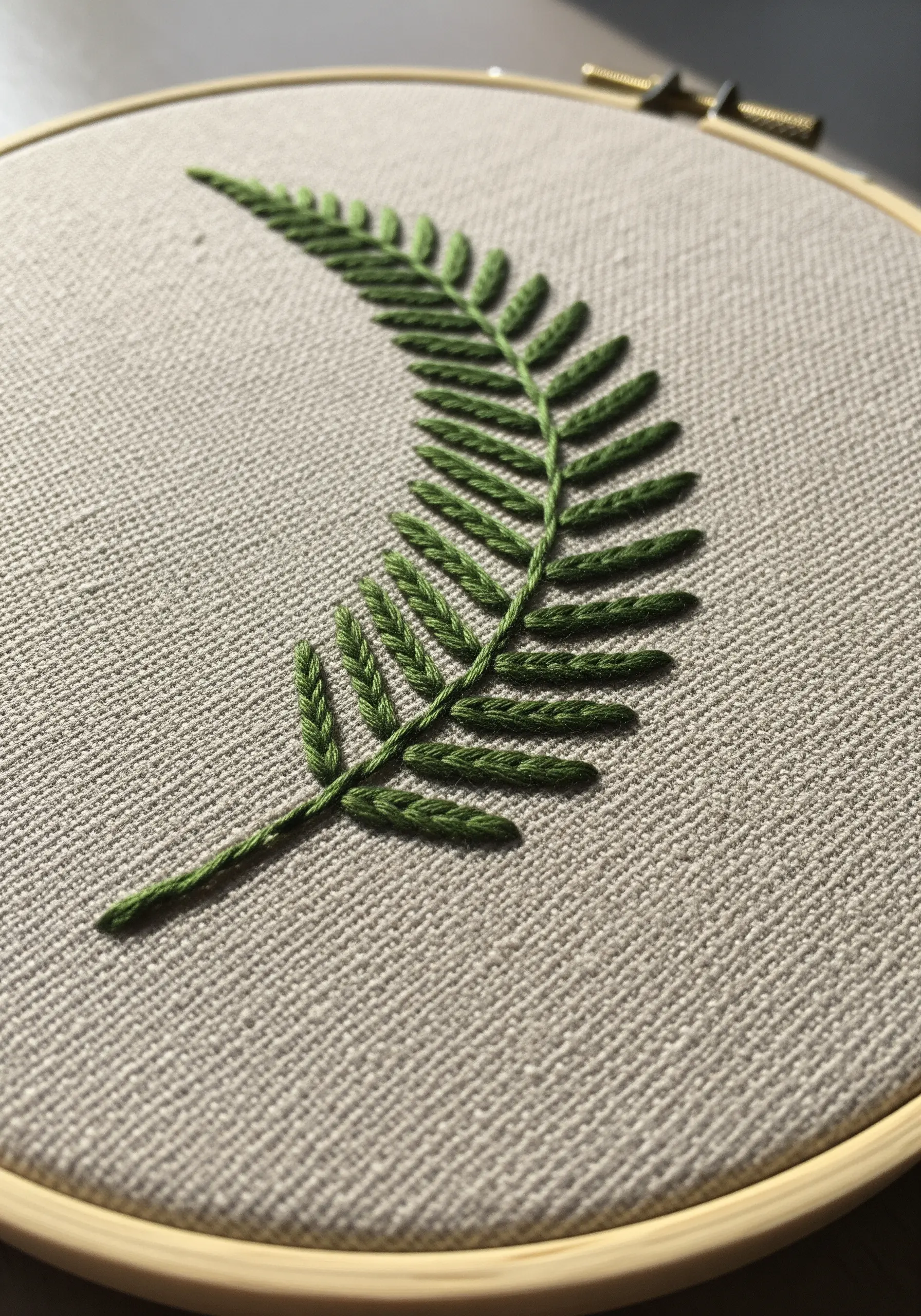 A single fern leaf embroidered in green thread using fishbone stitch on linen.