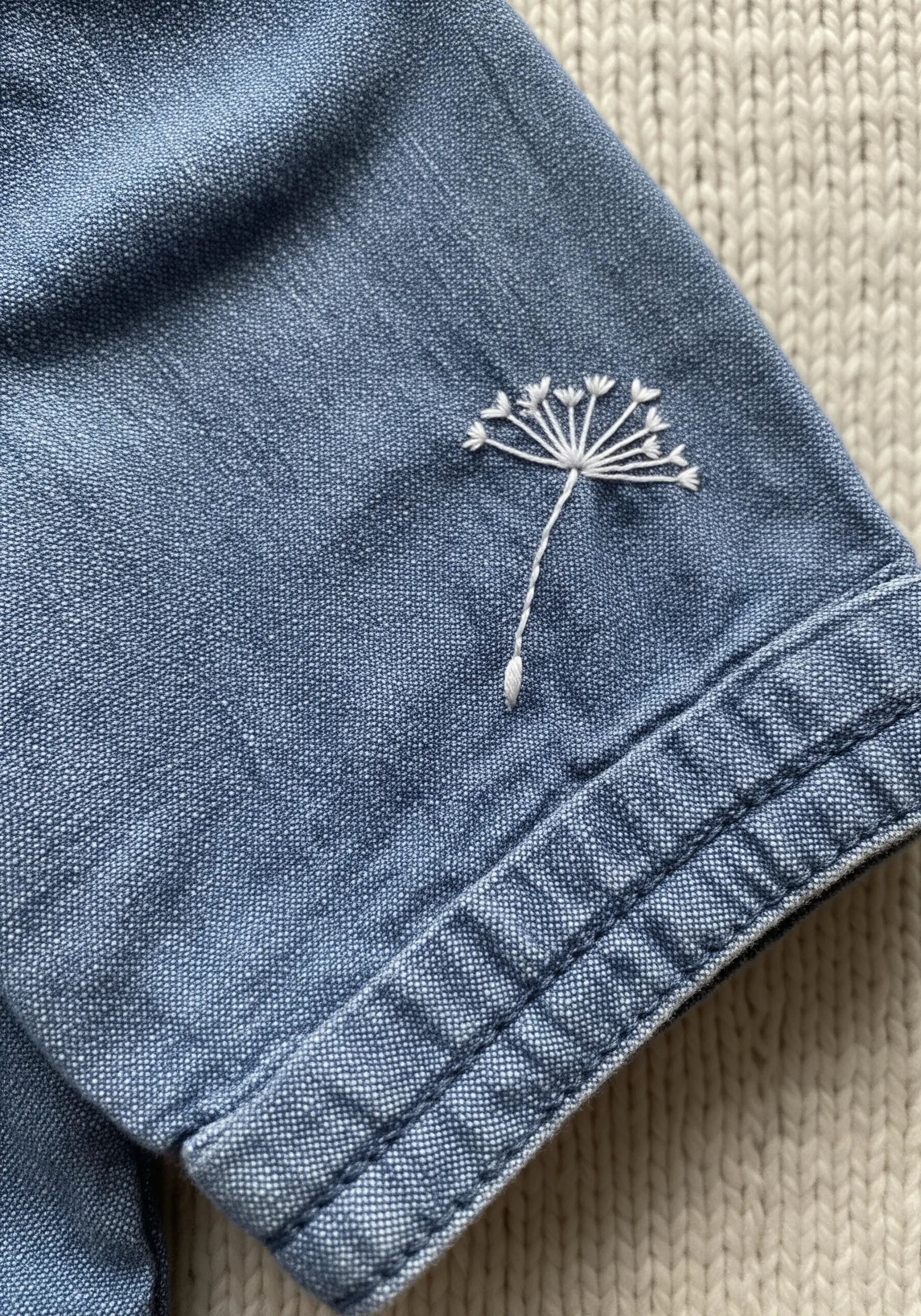 Delicate white dandelion seed head embroidered on a blue denim sleeve cuff.