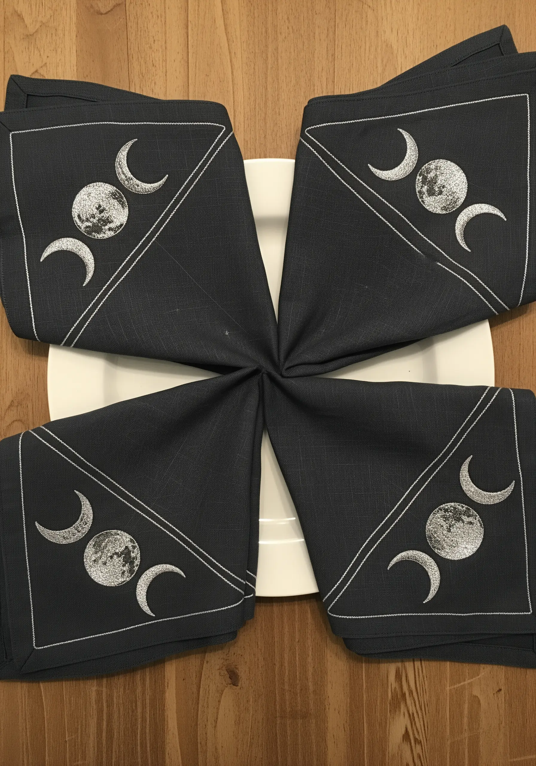 Four dark grey cloth napkins, each embroidered with a silver moon phase design.