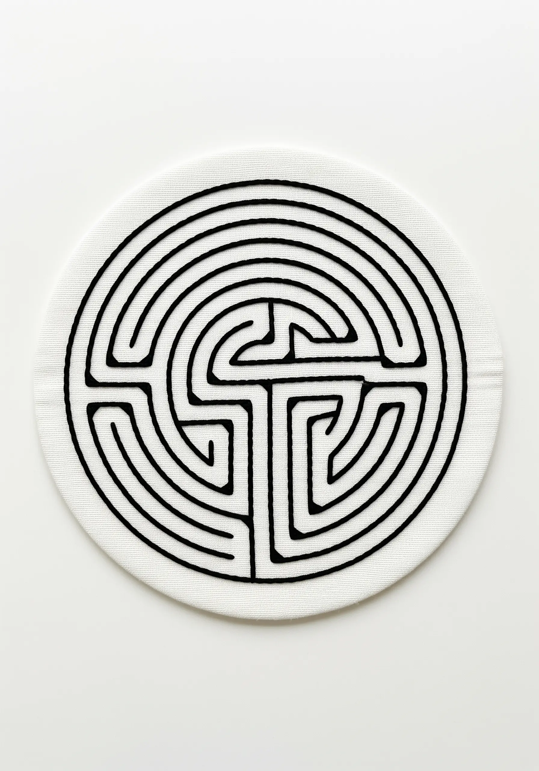 A circular labyrinth pattern stitched with thick, solid black embroidery on a white coaster.