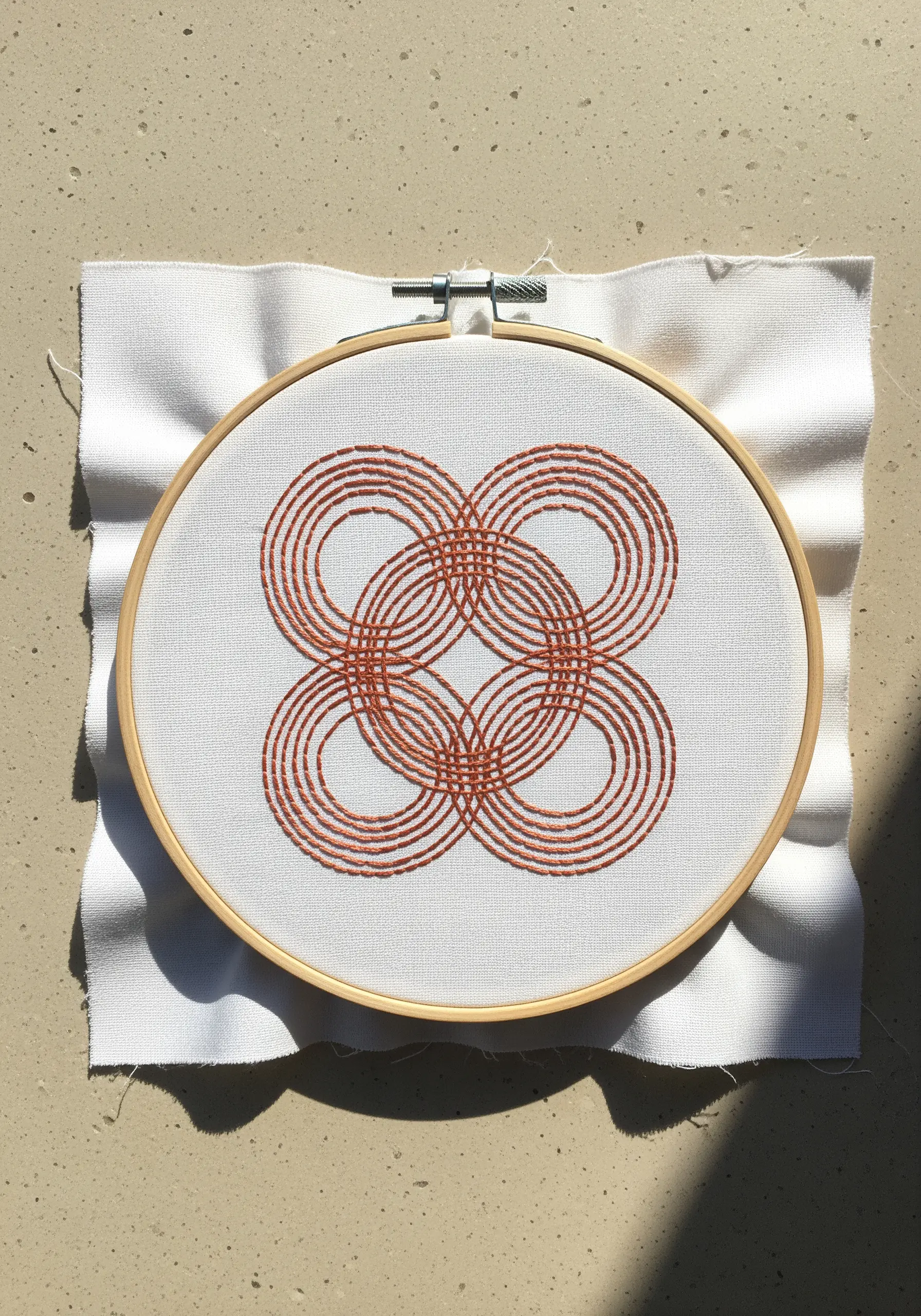 An intricate geometric knot of overlapping circles embroidered in orange thread on white fabric.