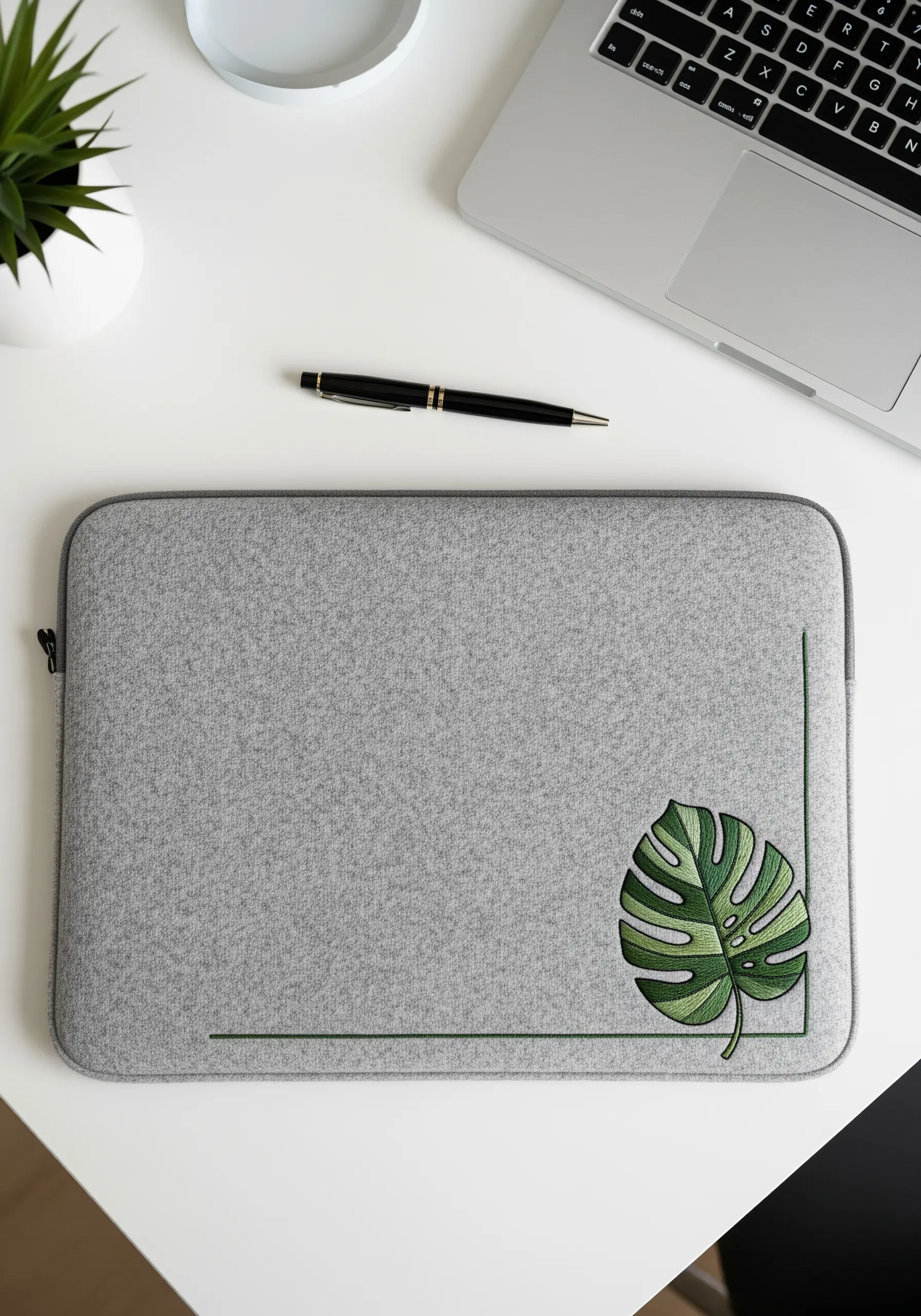 A grey felt laptop sleeve with a single, detailed green monstera leaf embroidered in the corner.
