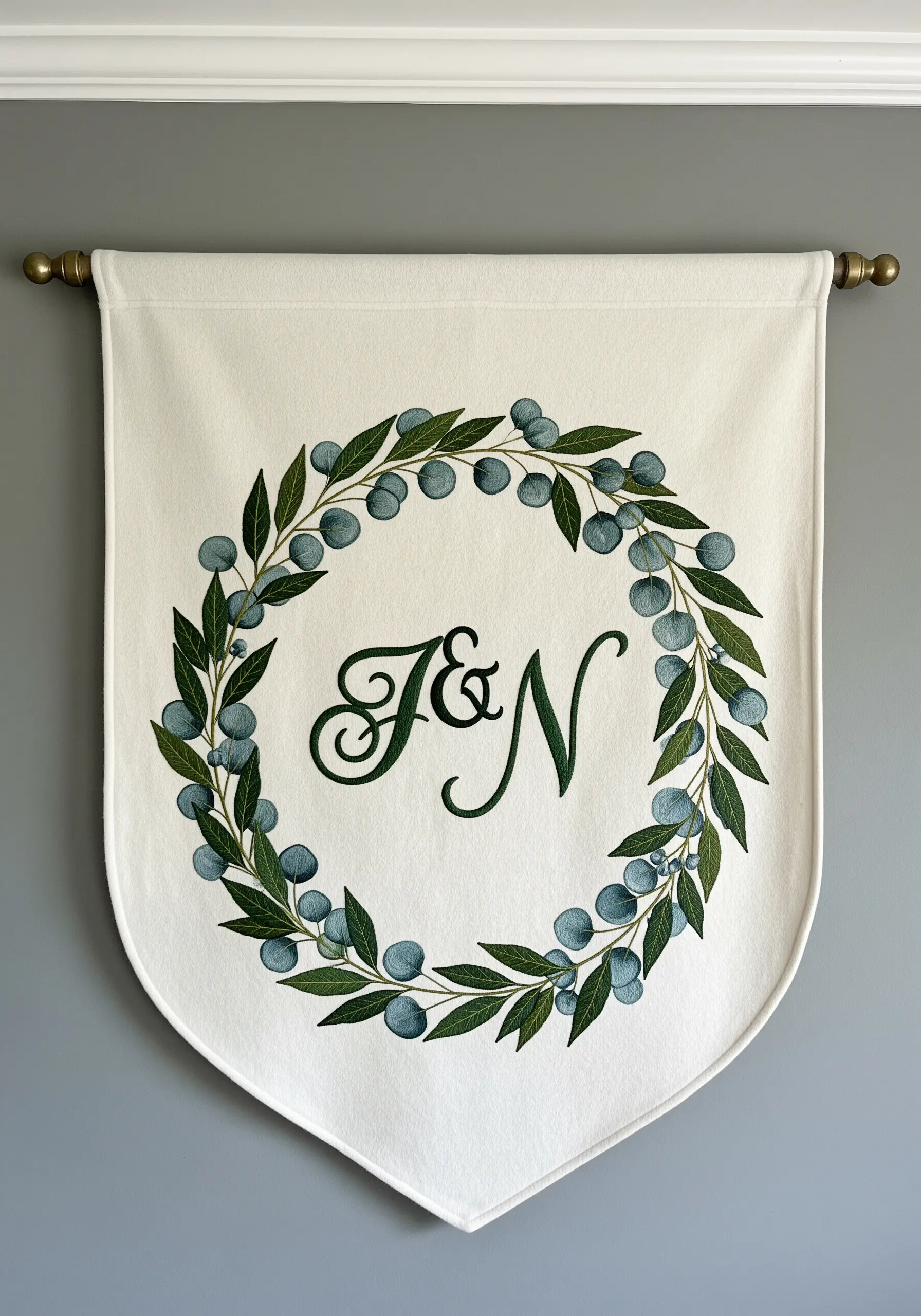 A white banner with a family monogram inside an embroidered wreath of green leaves and blue berries.