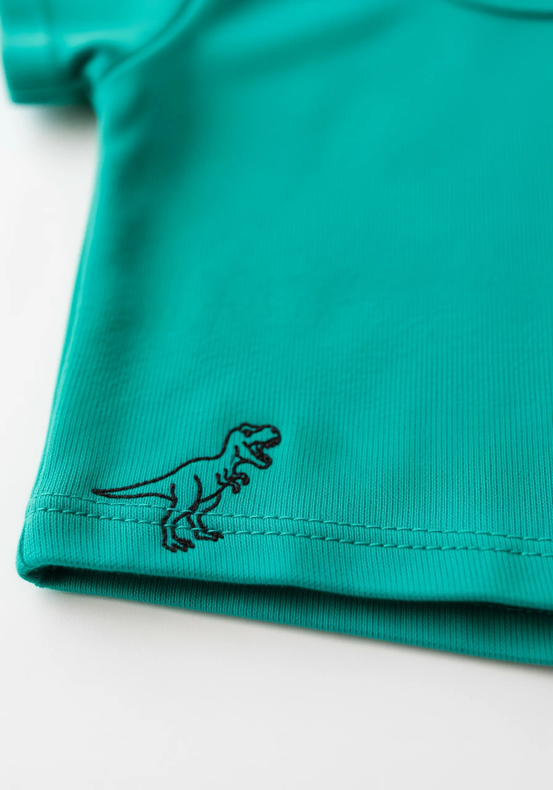 A small black line-art T-rex dinosaur embroidered on the hem of a teal shirt.