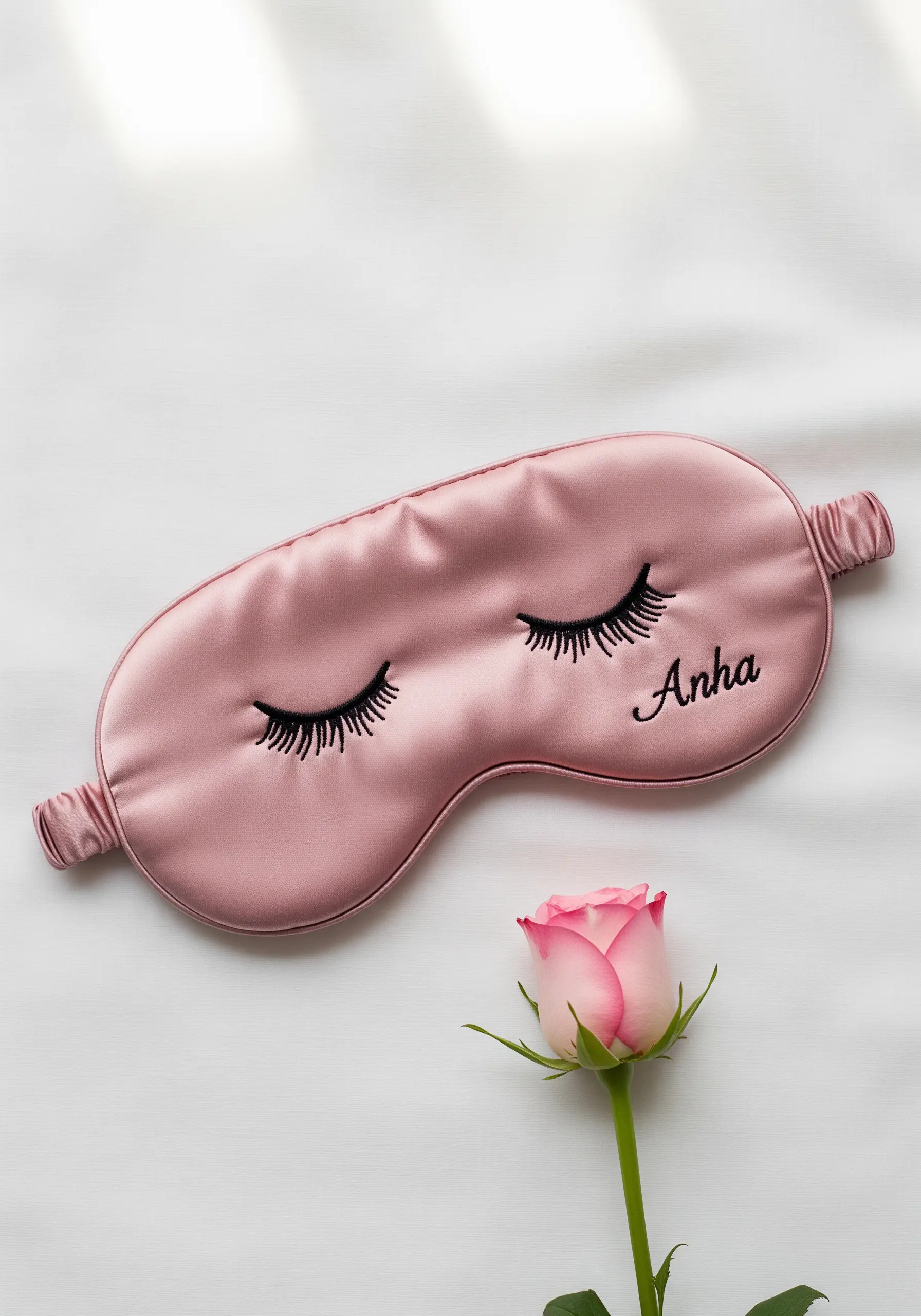 A pink satin sleep mask embroidered with a pair of closed eyes with long, dark eyelashes.