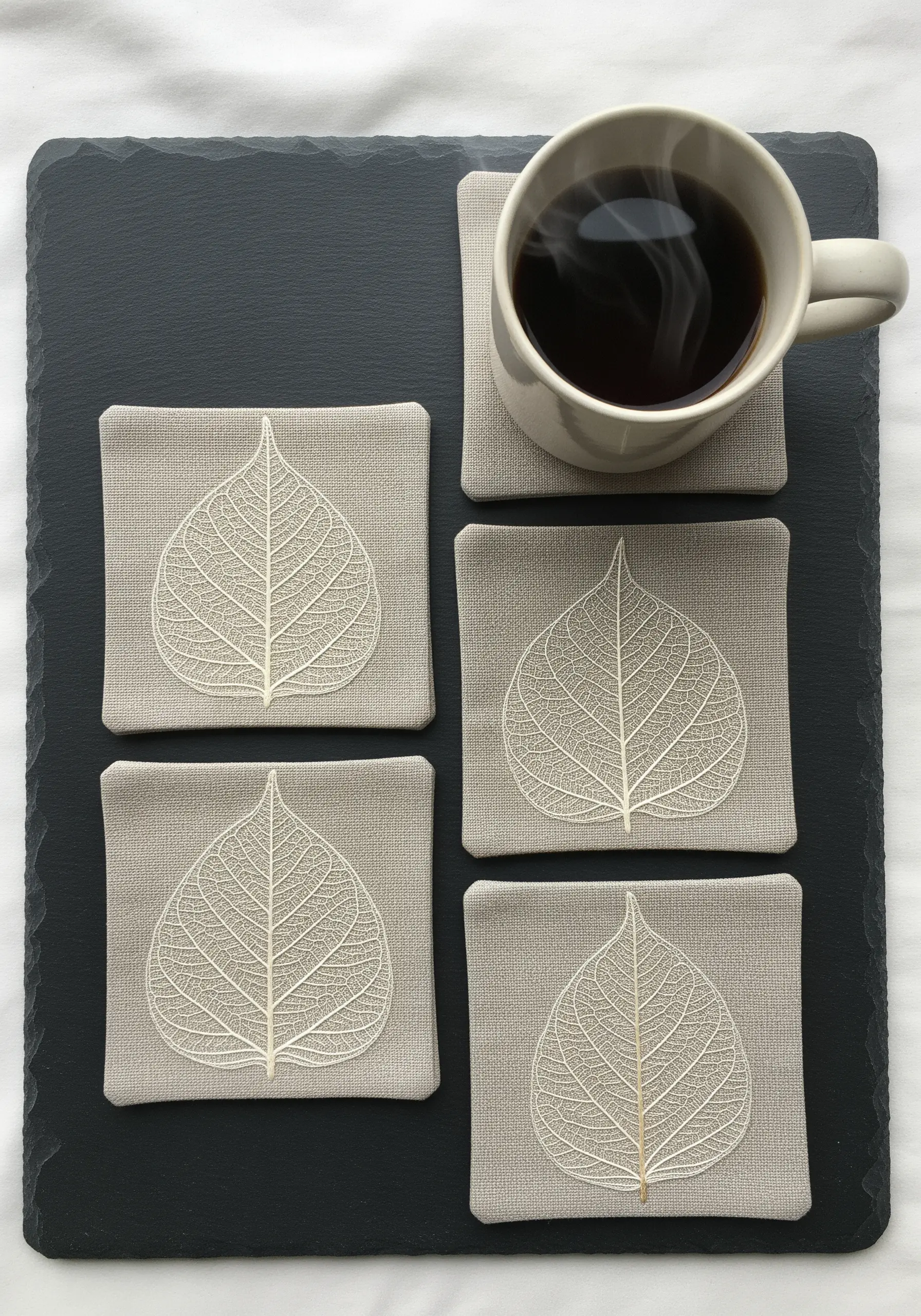 A set of four coasters, each embroidered with a delicate, white skeleton leaf on grey fabric.