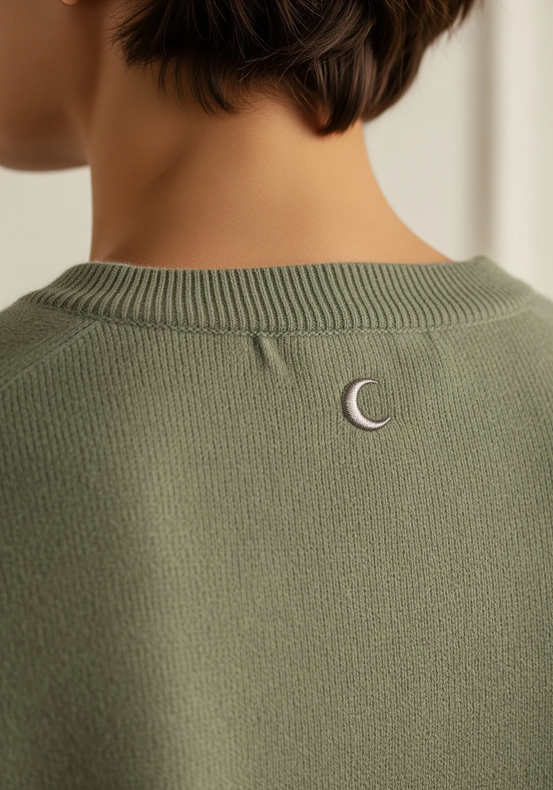 Small silver crescent moon embroidered on the back of a sage green sweater.