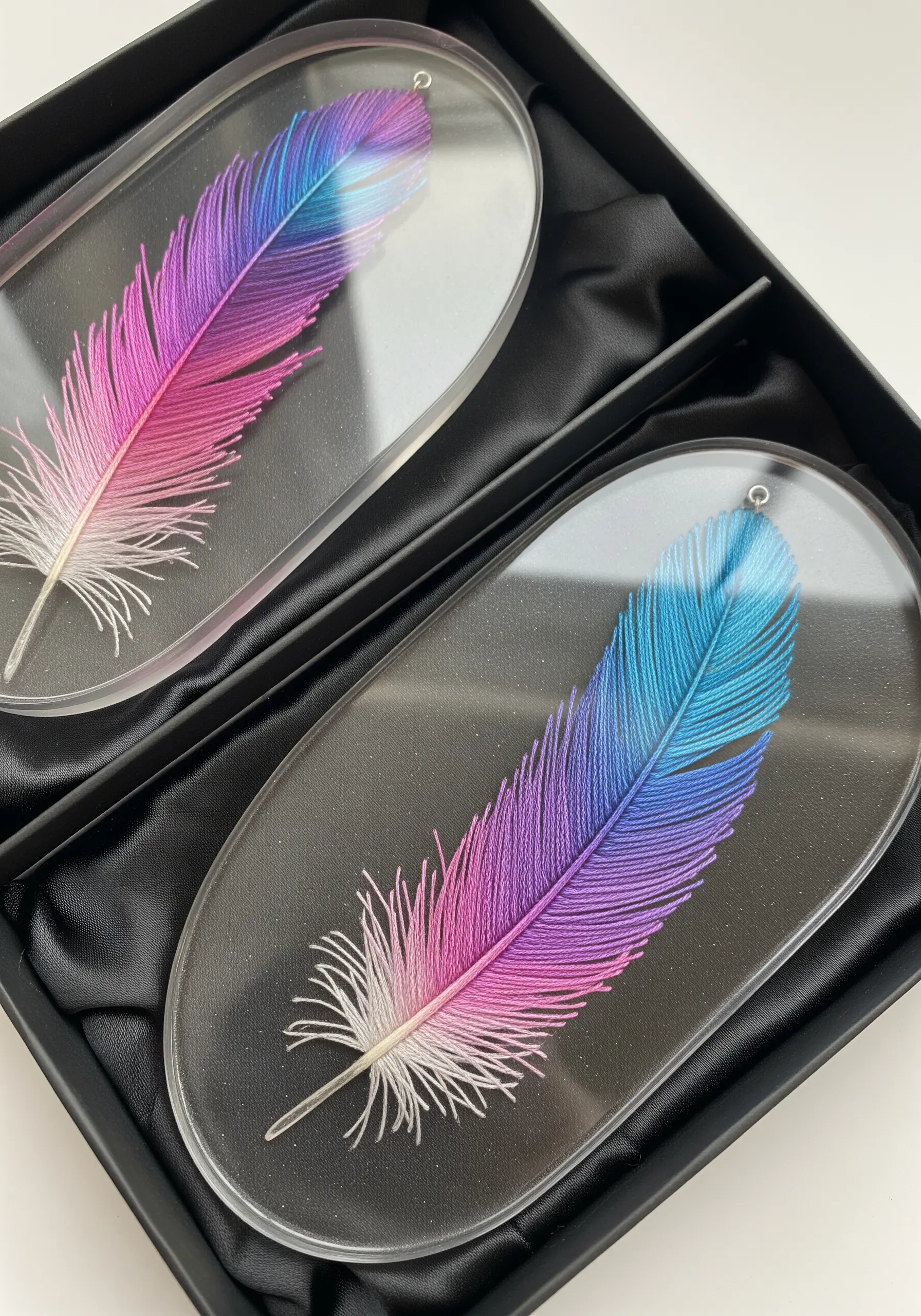 Two thread-painted feathers with pink, purple, and blue gradients sealed in clear oval resin tags.