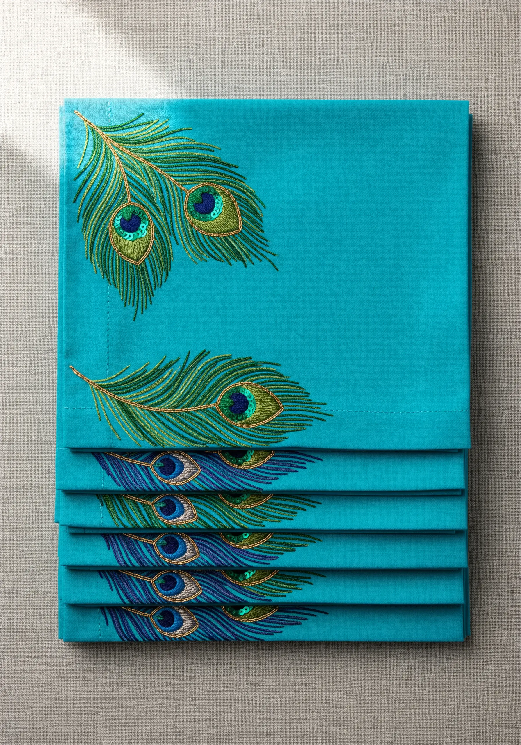 Embroidered peacock feathers with sequin details on a stack of turquoise napkins.