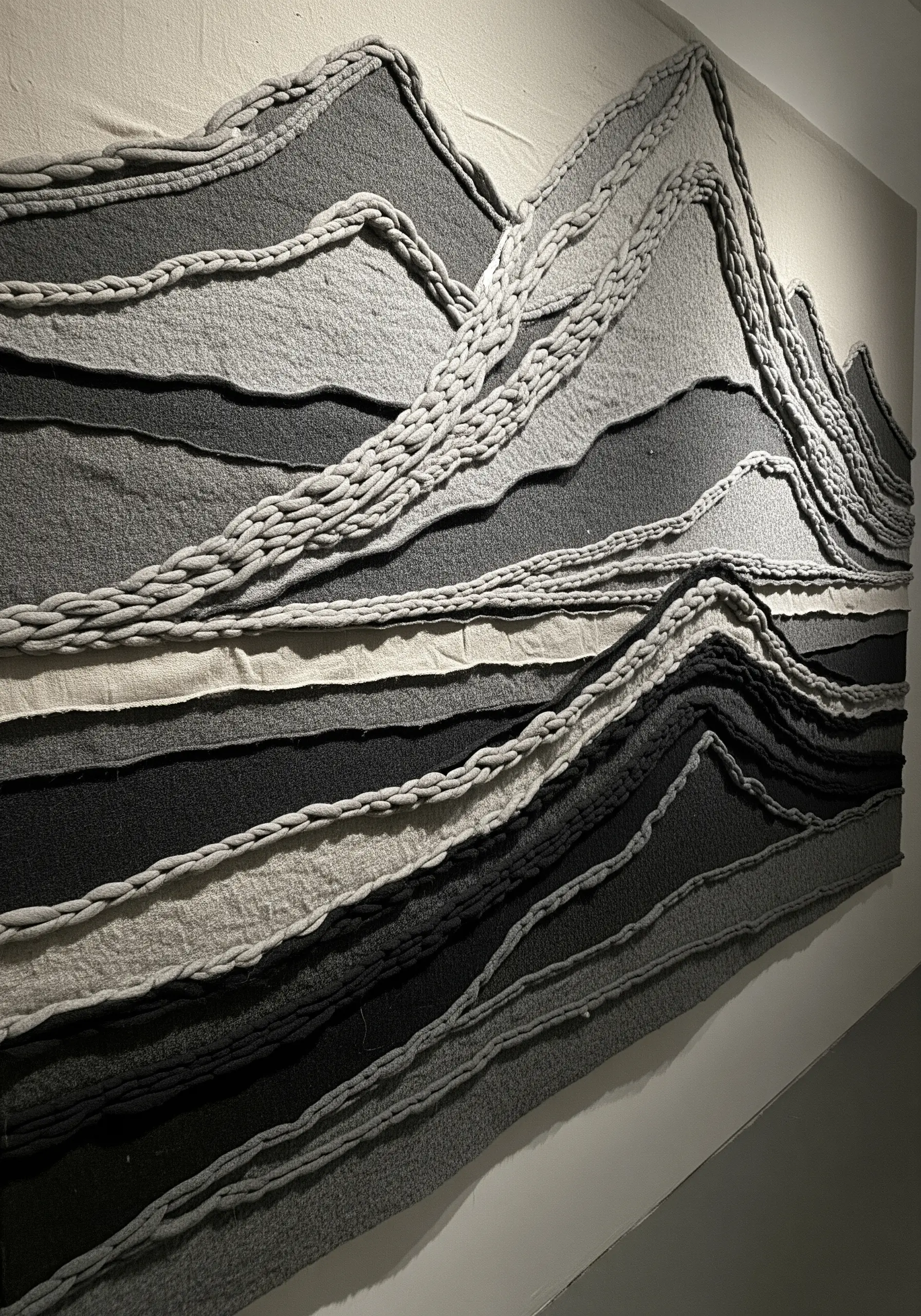 Monochrome textile wall art of mountains made from layered grey fabrics and thick braided cords.